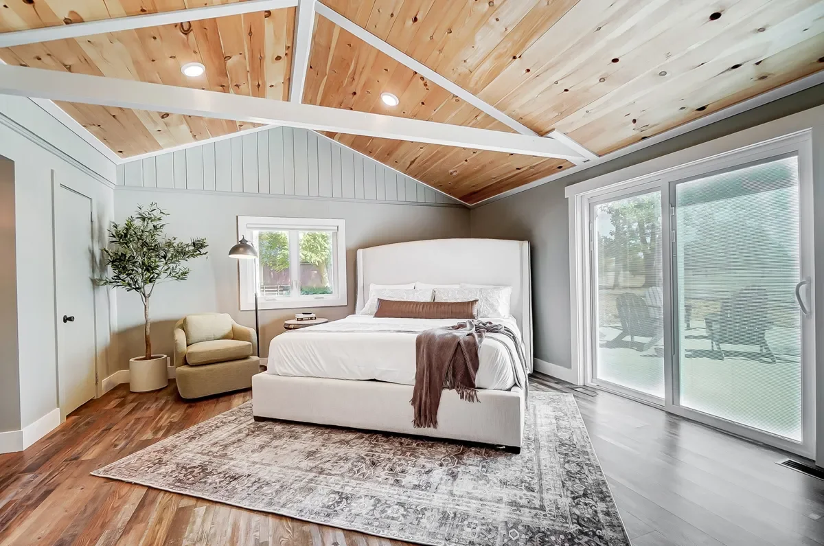 Bedroom with a vaulted wood ceiling, white walls, and a large sliding glass door leading to an outdoor patio. There is a white bed with a brown pillows and a throw blanket, a small window, a beige armchair with a floor lamp, a potted plant, and a pat