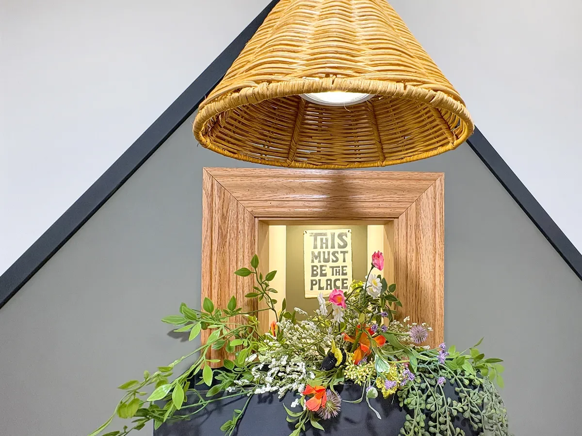 A decorative space with a triangular gray wall and black trim, featuring a wicker hanging lamp, a wooden-framed mirror, and a floral arrangement with various green, pink, orange, purple, and white flowers and foliage below the mirror. In the mirror's