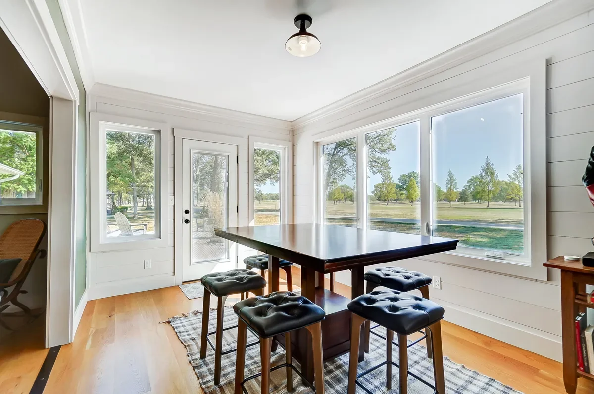 Sunlit dining area with large windows and a wooden table surrounded by six black upholstered stools, overlooking a grassy outdoor landscape.