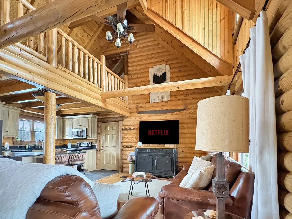 Interior of a log cabin living room with wooden walls and ceiling, a leather sofa with throw pillows, a black television on a black cabinet displaying Netflix, a floor lamp, a small coffee table, and an open kitchen area in the background. There are 