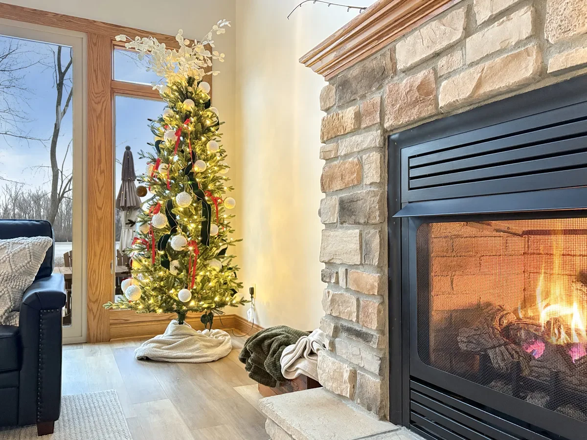 A decorated Christmas tree with white ornaments and red ribbons in a living room next to a fireplace with a fire burning.