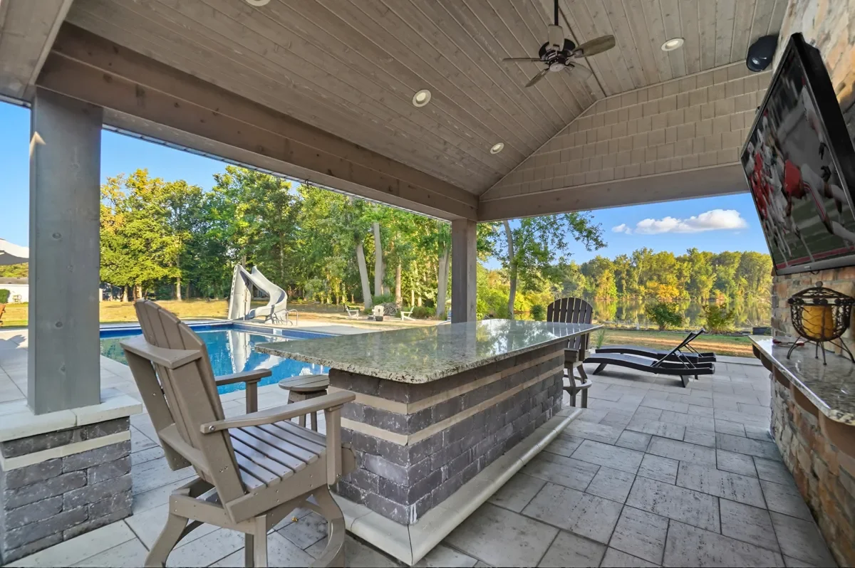 Covered patio with a pool, water slide, lounge chair, chairs around a table, and scenic view of trees and water.