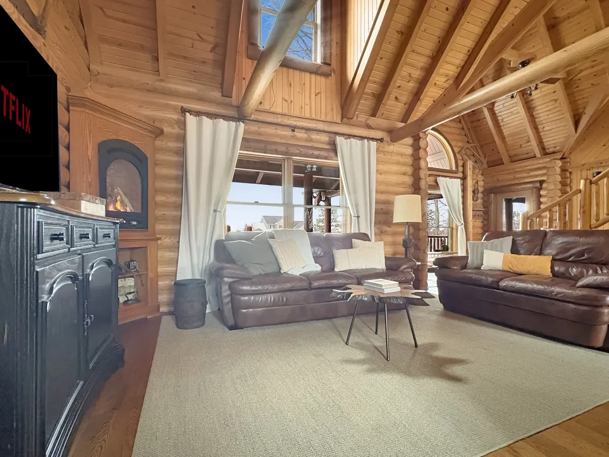 A cozy living room in a log cabin with wooden walls and ceiling, featuring two leather sofas with throw pillows, a small black coffee table with books, a floor lamp, a window with white curtains, and a black fireplace or cabinet on the left.