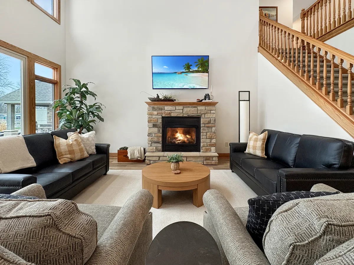 A living room with two black sofas, a beige armchair, a round wooden coffee table with a small potted plant, a fireplace with a stone surround, a wall-mounted TV displaying a tropical beach scene, a large window with blinds, and a staircase with wood