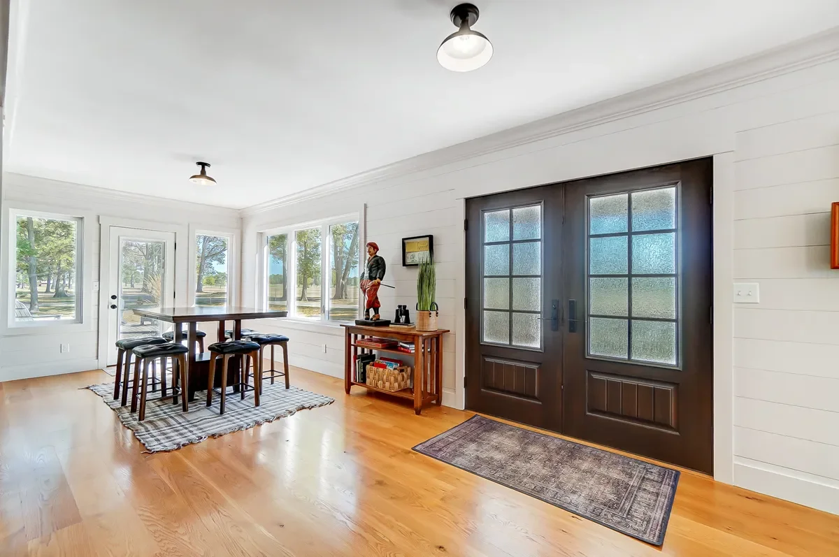 Bright living room with white walls, hardwood floors, large windows, and a dark double door with frosted glass panes. A wooden side table with decor and a small rug are near the door, and a dining table with stools is placed on a larger rug.