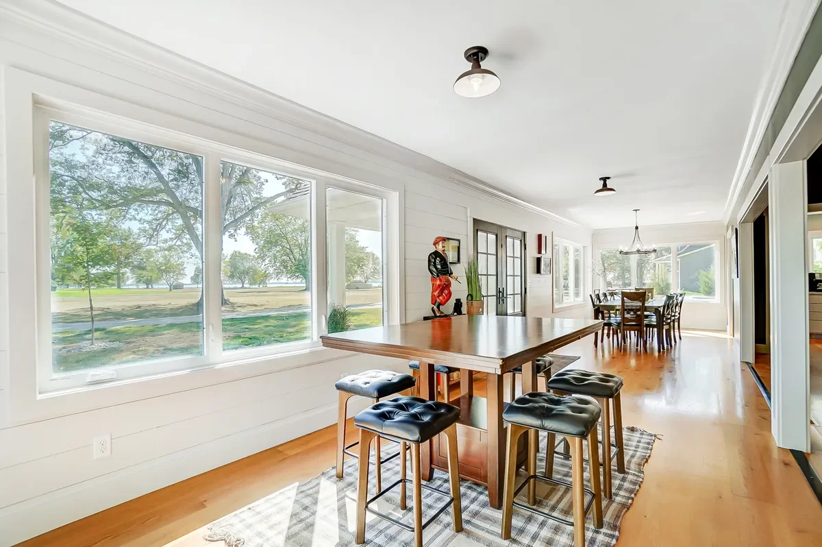 Bright dining room with large windows overlooking a park and trees, containing a wooden table with four black cushioned stools, a tasteful chandelier, and a sideboard with decorative items.
