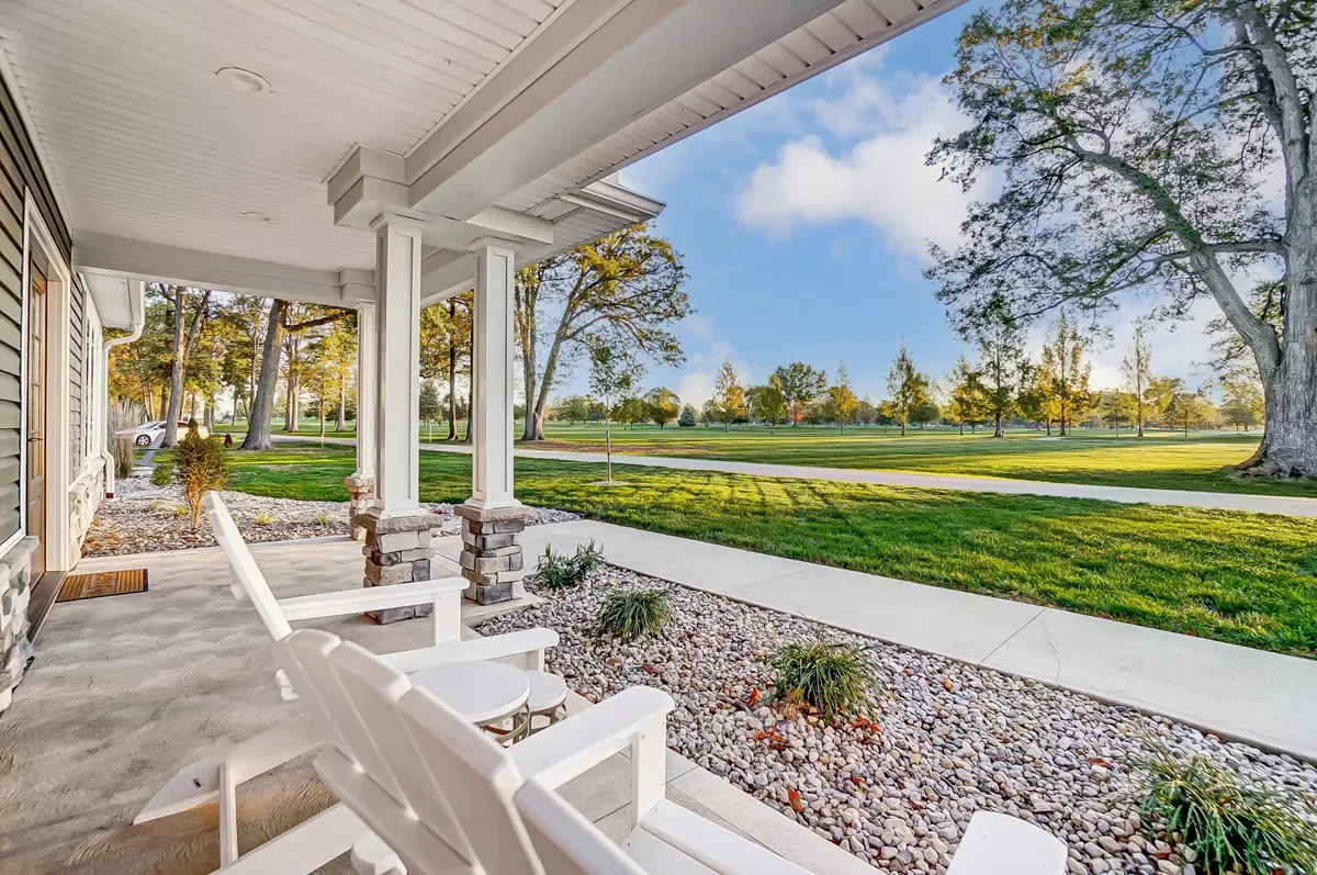 Front porch of a house with white chairs, brick pillars, and a view of a green park with trees under a partly cloudy sky.