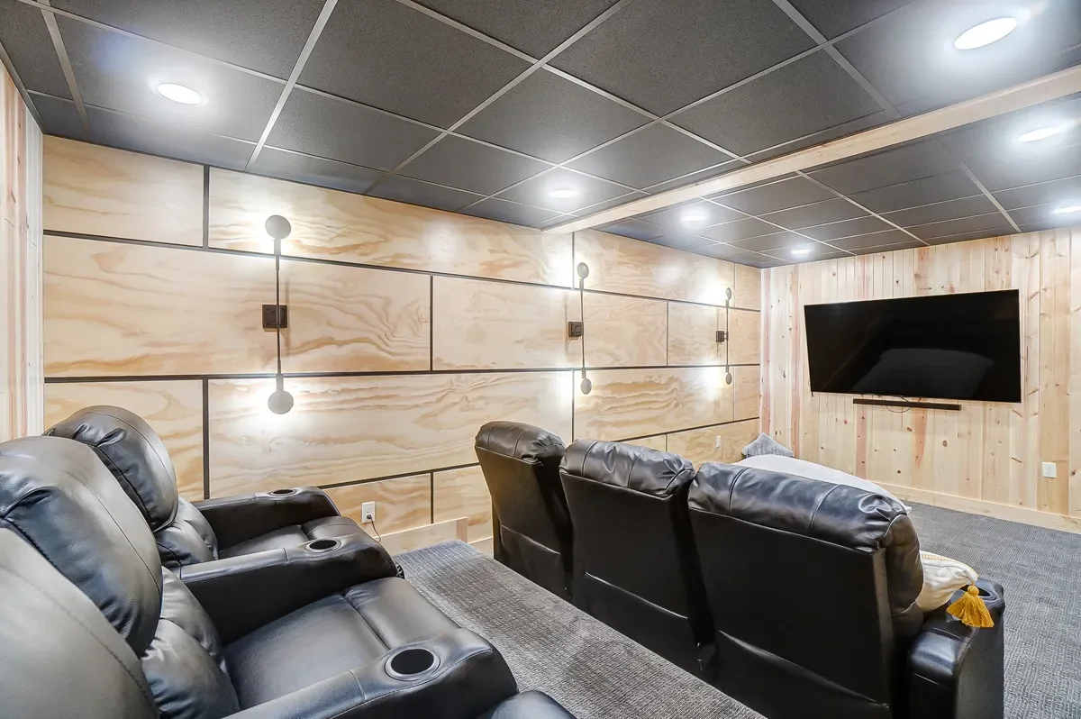 Home theater room with black leather recliners, a large flat-screen TV on a wooden-paneled wall, and modern wall-mounted light fixtures.