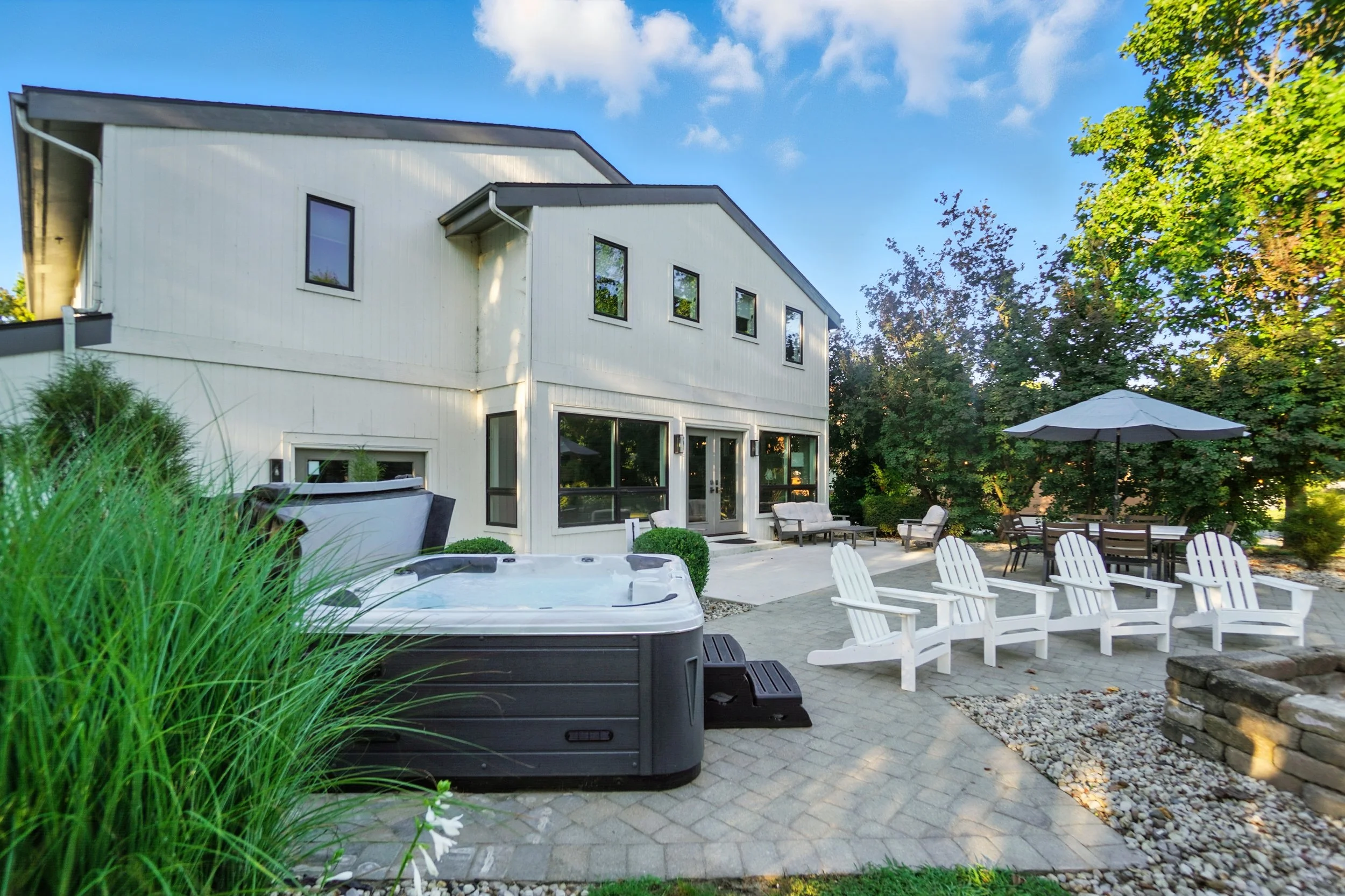 Backyard patio with a hot tub, white Adirondack chairs, a table with an umbrella, and outdoor seating area near a modern two-story house with multiple windows, surrounded by trees under a blue sky.