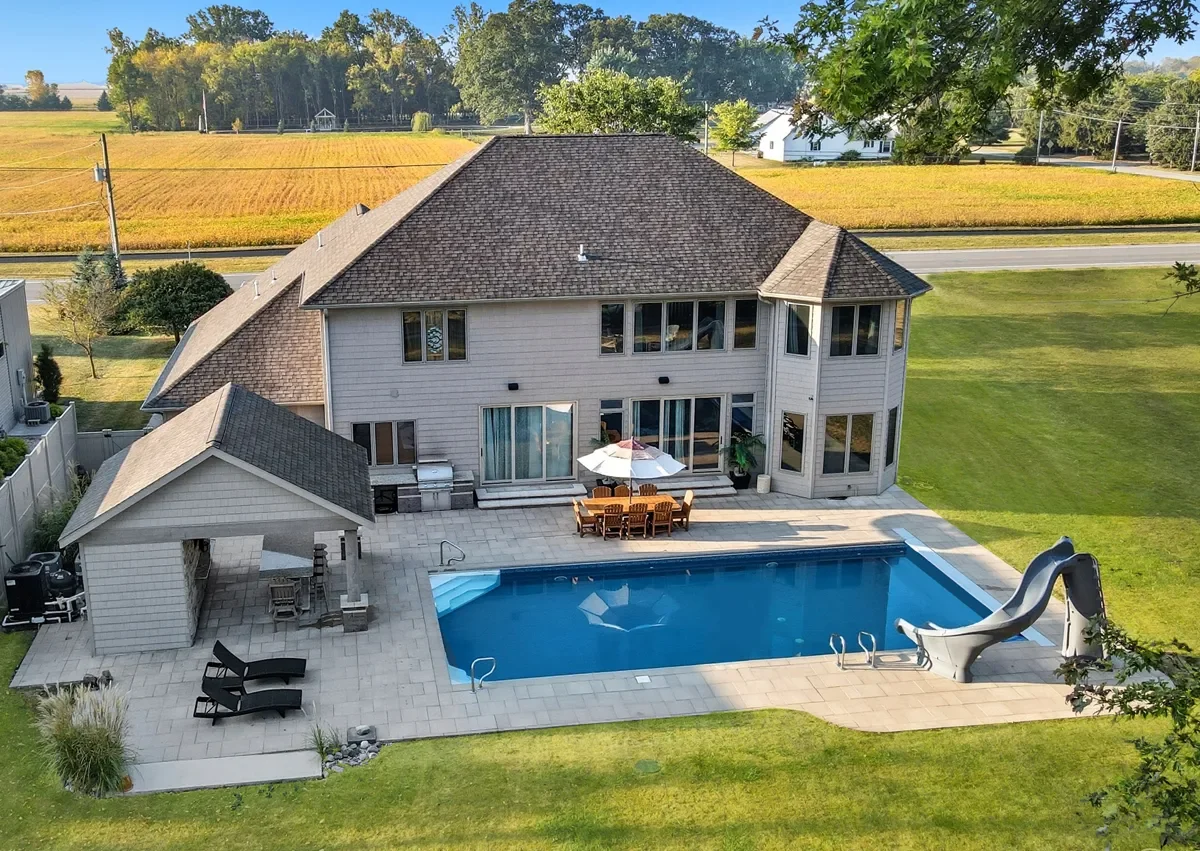 A backyard view of a modern house with a swimming pool, slide, outdoor dining area with an umbrella, lounge chairs, and a small covered sitting area, surrounded by a grassy lawn and fields.