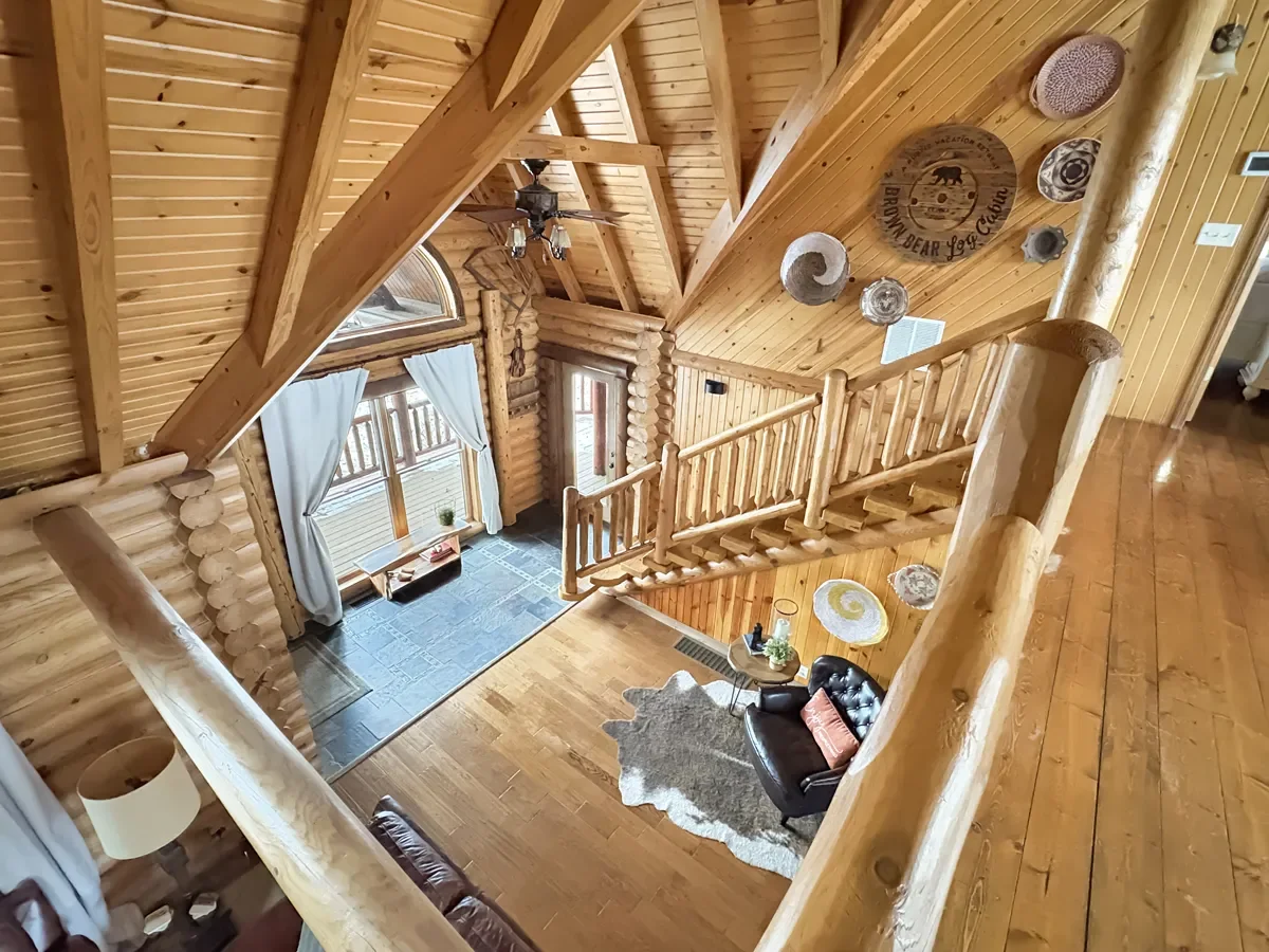 View of a log cabin interior showing a living room with wood-paneled walls and ceiling, a staircase railing, a black leather couch, a small side table with decor, a cowhide rug, and a large window with curtains. Wall decorations include circular art 