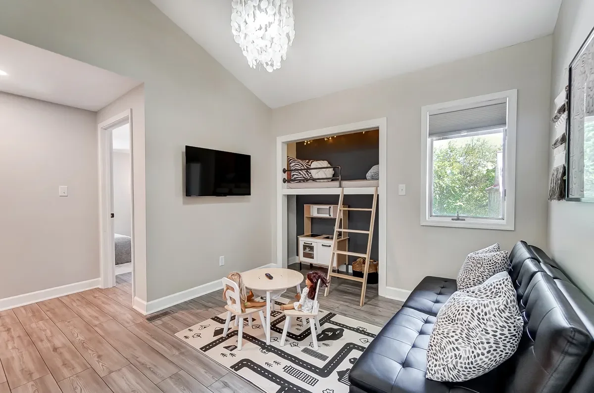 Living room with a black futon, leopard print pillows, small play table with dolls, wall-mounted TV, window showing greenery, and a loft bed with ladder and bedding.