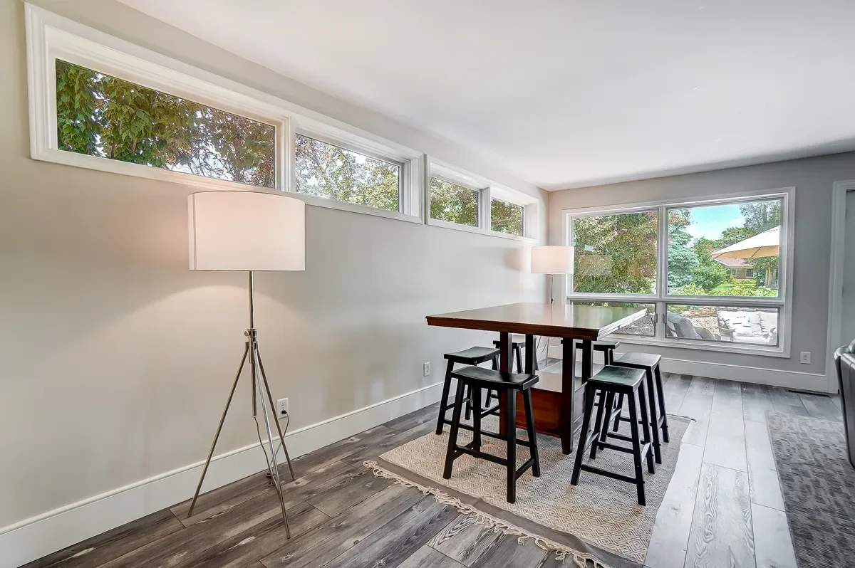 A dining area with a wooden table, four black stools, a tall standing lamp, and large windows with a view of a patio with outdoor furniture and trees.