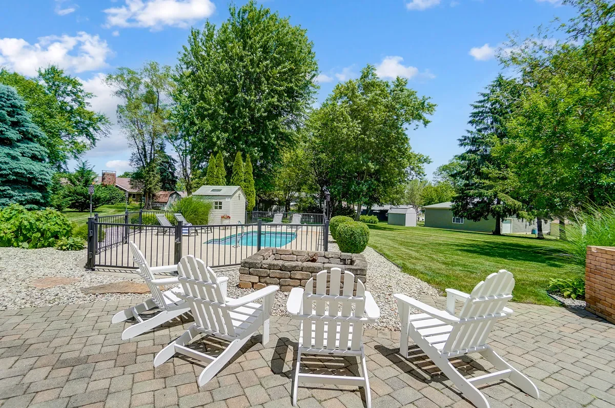 Backyard with a patio, white chairs, a fire pit, a fenced-in swimming pool, garden shed, lush green trees, grassy lawn, and a partly cloudy sky.