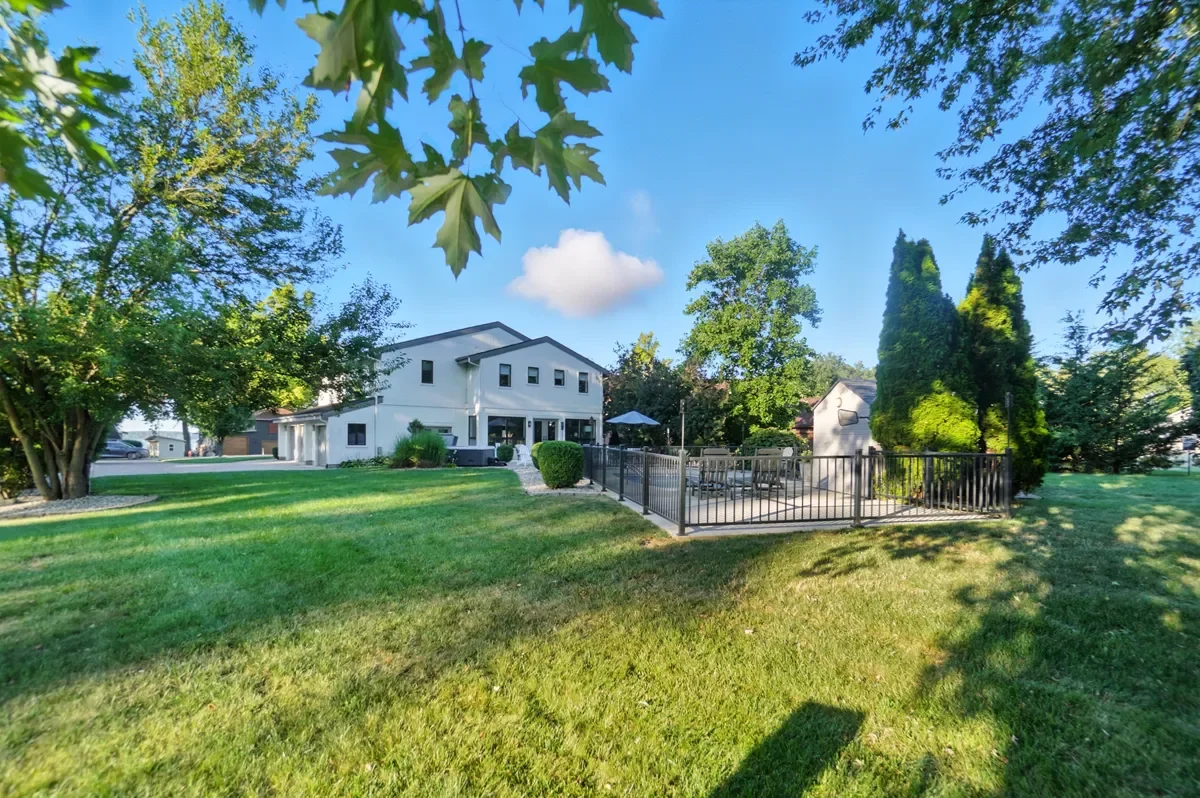 Backyard with a modern white house, a fenced patio area with outdoor furniture, surrounded by lush green trees and grass, under a blue sky with a few clouds.