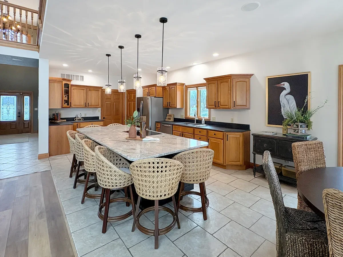 A spacious kitchen with wooden cabinets, a large island with a granite countertop, hanging pendant lights, and a black sideboard with plants and artwork on the wall.