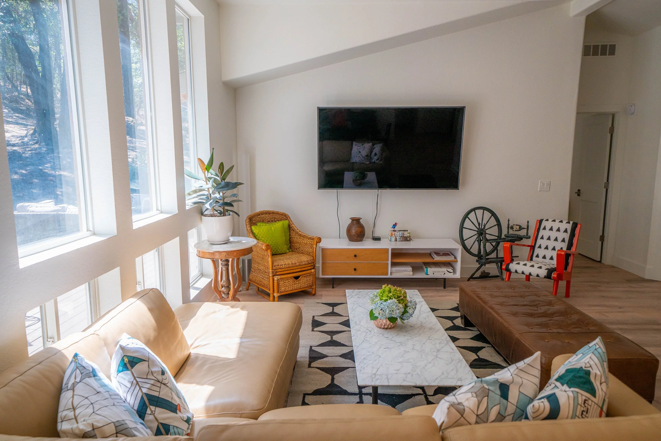 Modern living room with beige sofa, white marble coffee table, wall-mounted TV, patterned rug, wicker chair, potted plant, brown leather ottoman, and decorative pillows.