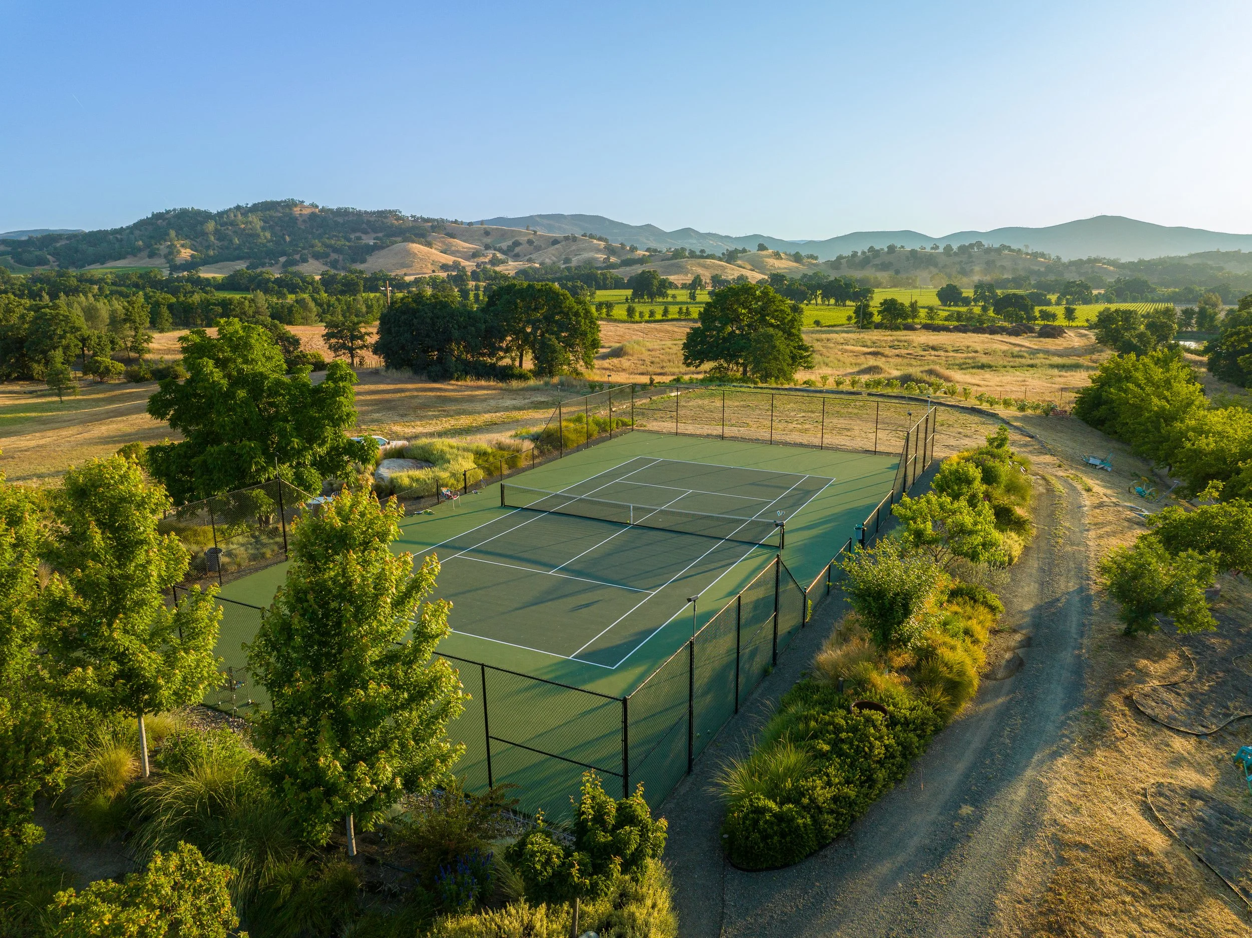 An outdoor tennis court surrounded by green trees and open countryside with rolling hills in the distance, under a clear blue sky.