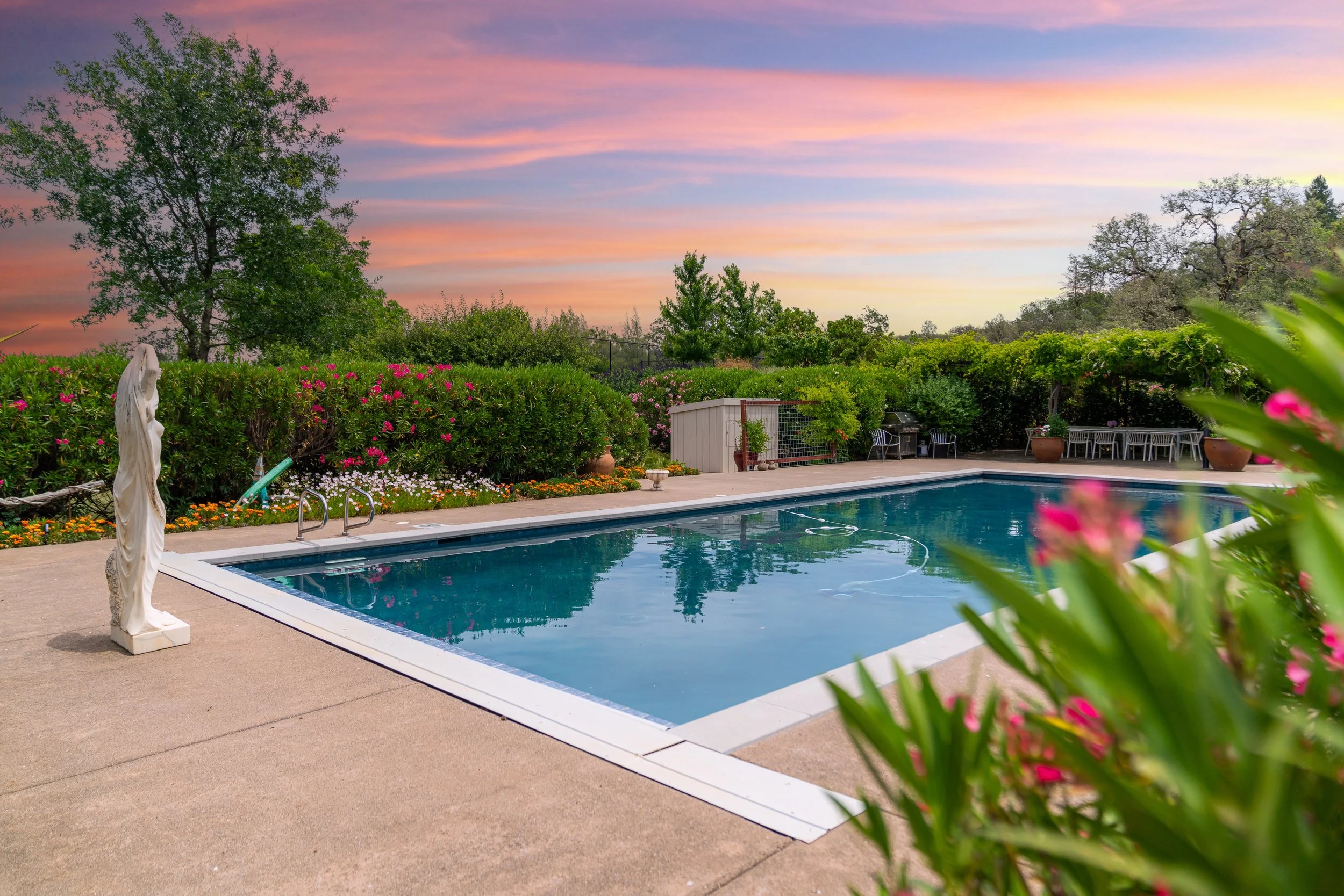An outdoor swimming pool surrounded by greenery and colorful flowers at sunset, with garden furniture and a statue of a woman near the pool.