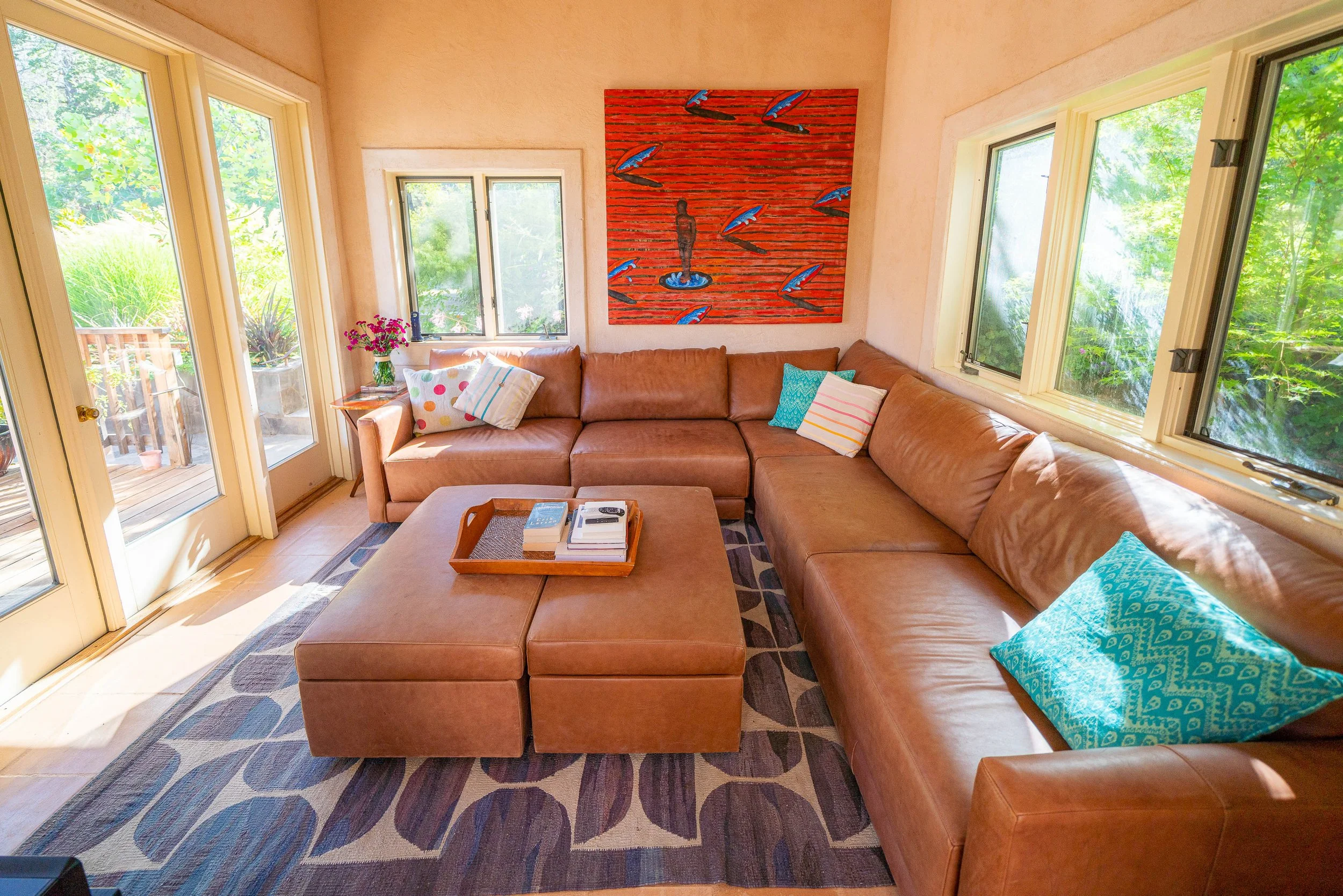 Living room with a large tan leather sectional sofa, colorful pillows, a patterned rug, a glass door leading outside, windows with green foliage, and a red and blue abstract artwork on the wall.