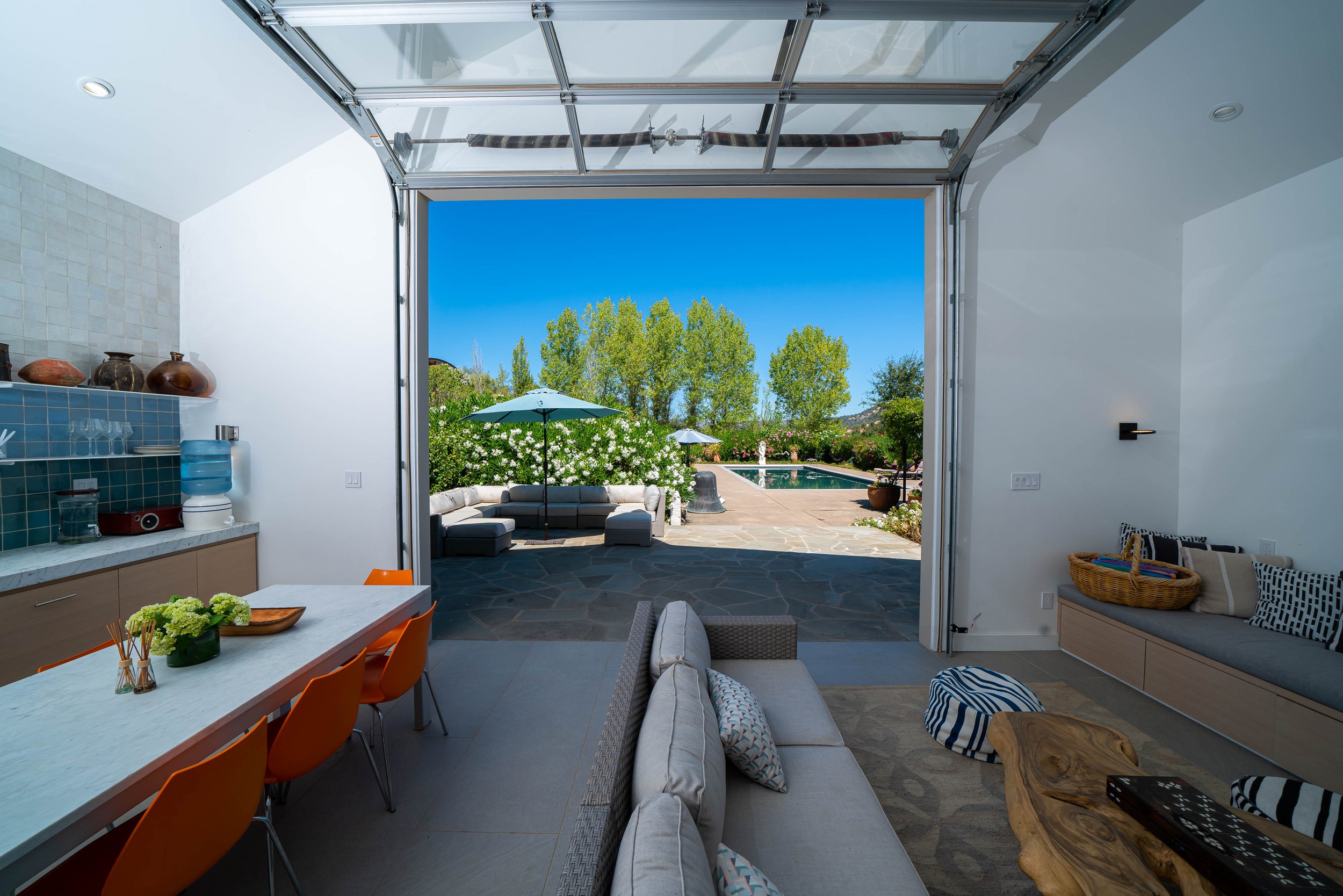 View from inside a modern living room looking out to a backyard with patio, pool, trees, and clear blue sky.