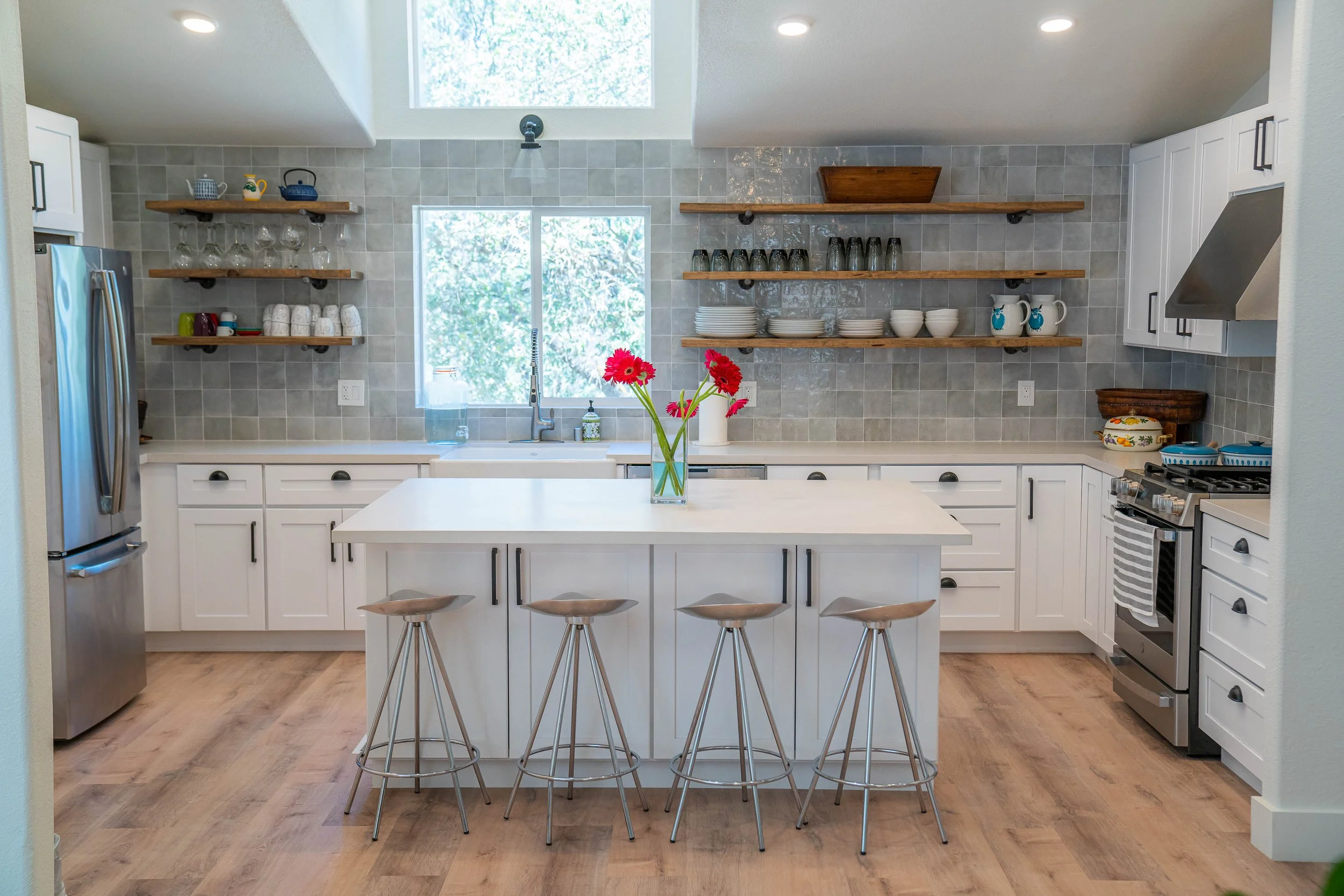 Modern kitchen with white cabinets, a central island with four bar stools, open wooden shelves with dishes and glassware, a window above the sink, and a stainless steel refrigerator.
