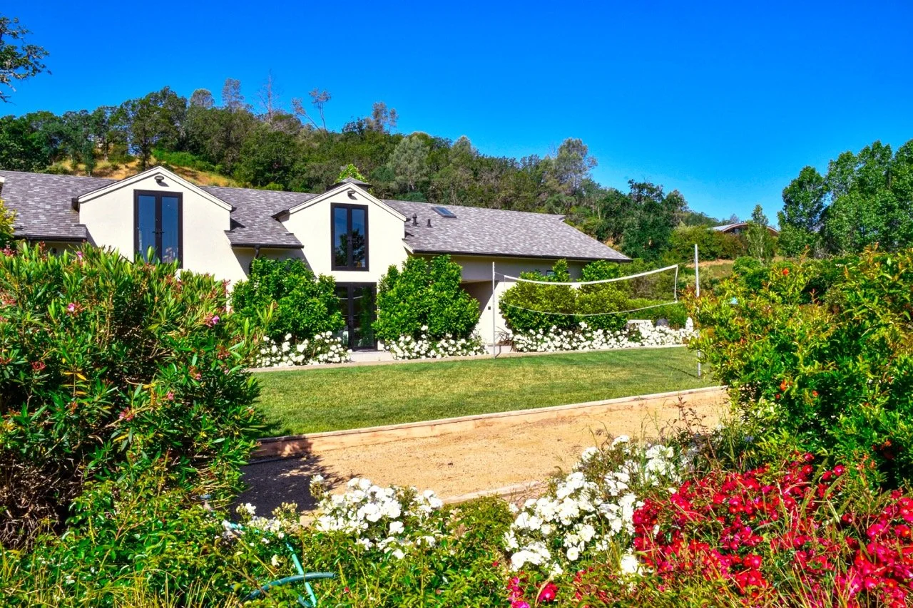 A modern white house with large windows, surrounded by greenery and colorful flowers, under a bright blue sky.