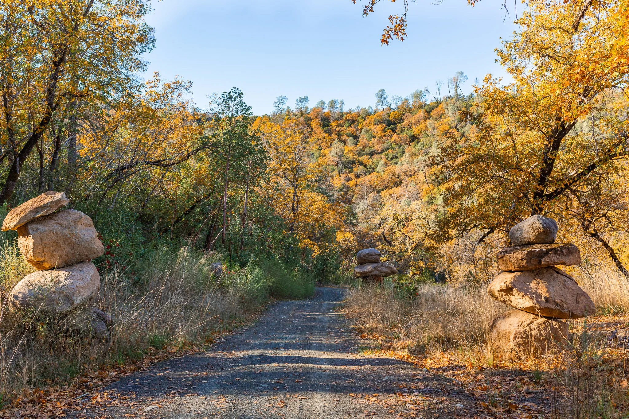 A dirt road flanked by stacked rocks on both sides, with autumn-colored trees and hills in the background.