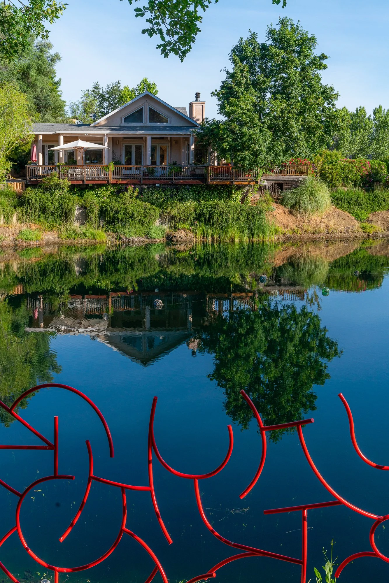 Residential house with deck along a calm body of water, trees and bushes surrounding it, and a red metal sculpture in the foreground with reflections on the water.