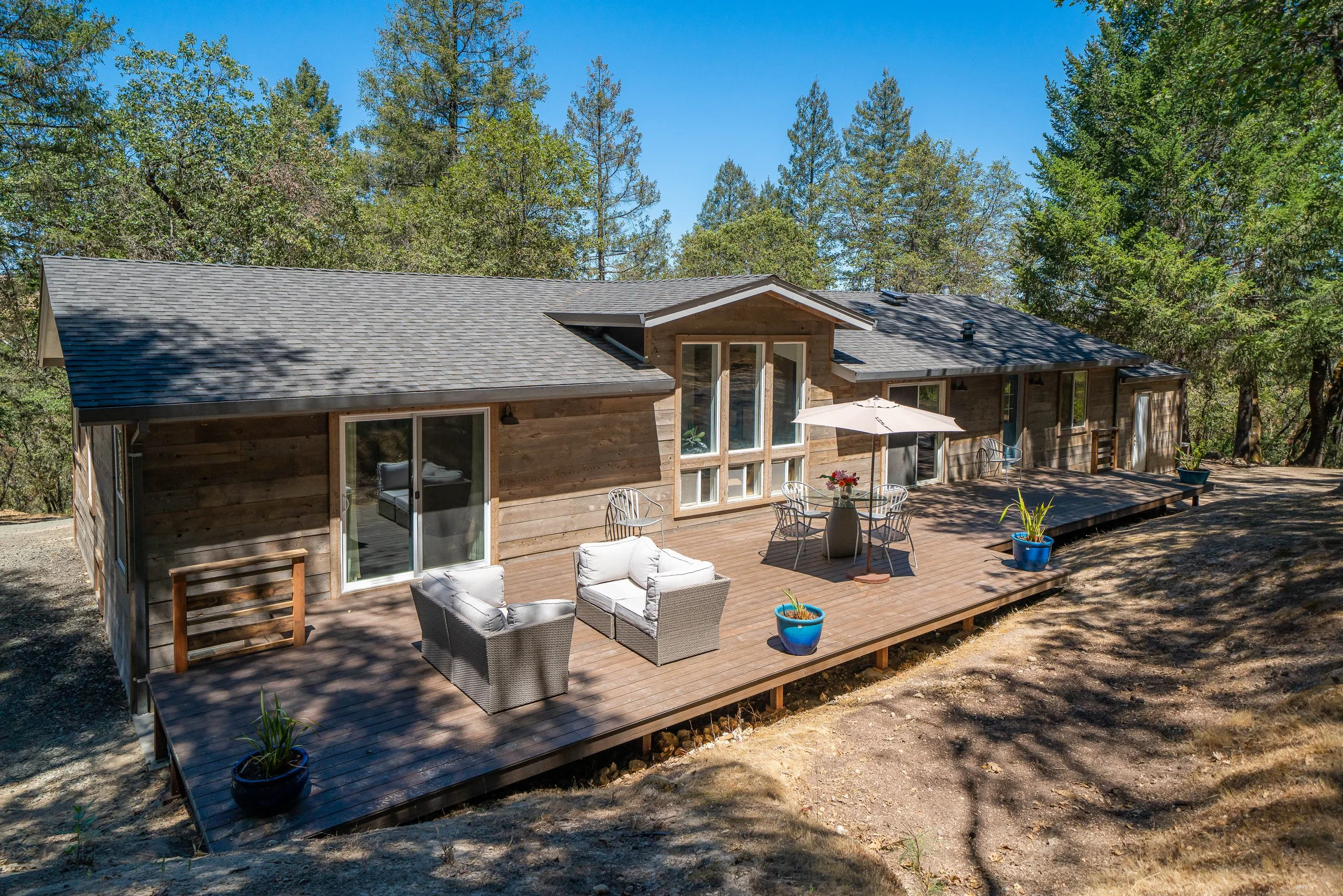 Wooden house with a large deck, outdoor seating, potted plants, and surrounded by trees.