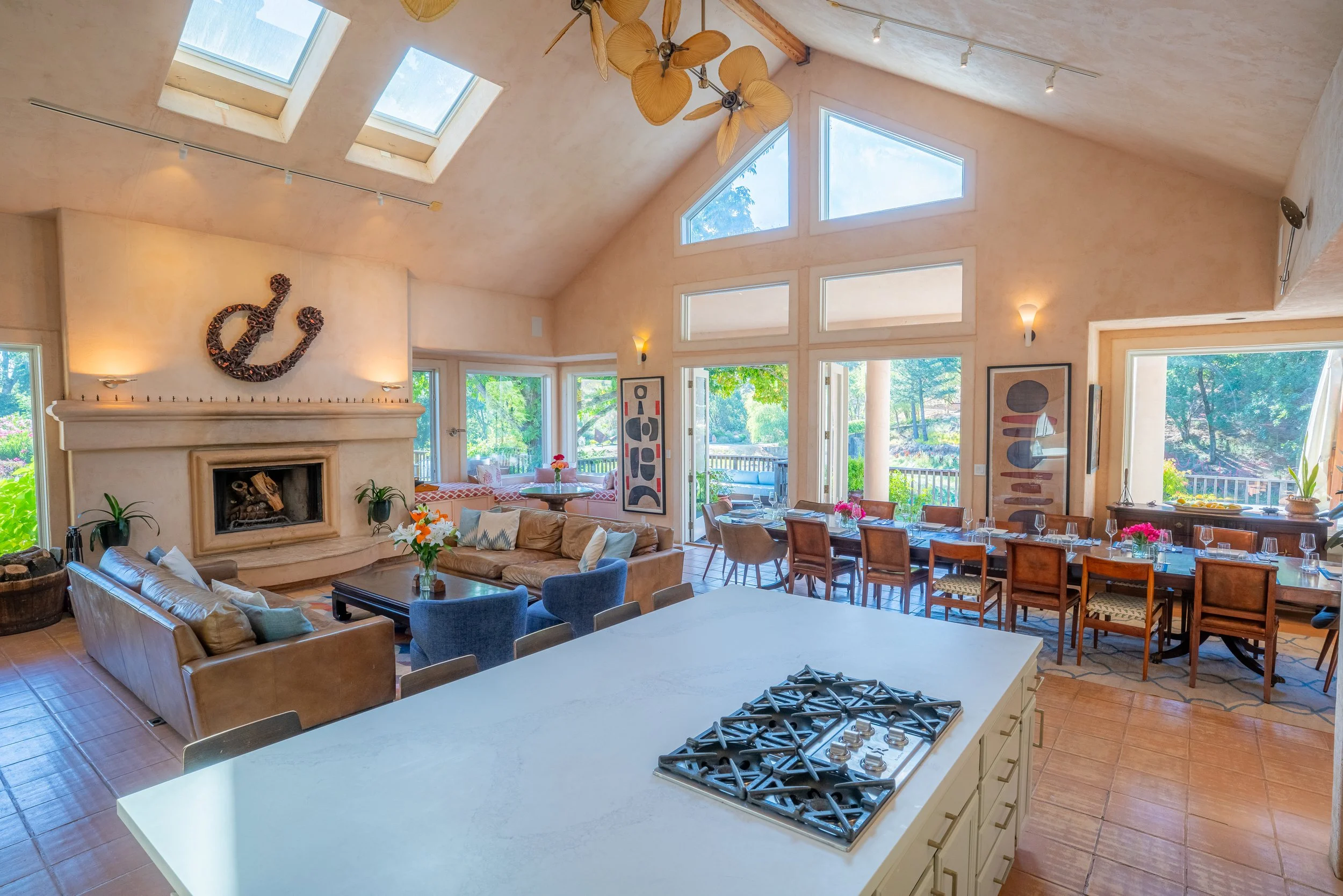 Open-concept living and dining area with large windows and skylights, featuring a kitchen island with a gas stove, a fireplace, colorful artwork, and various seating arrangements in a bright, modern space.