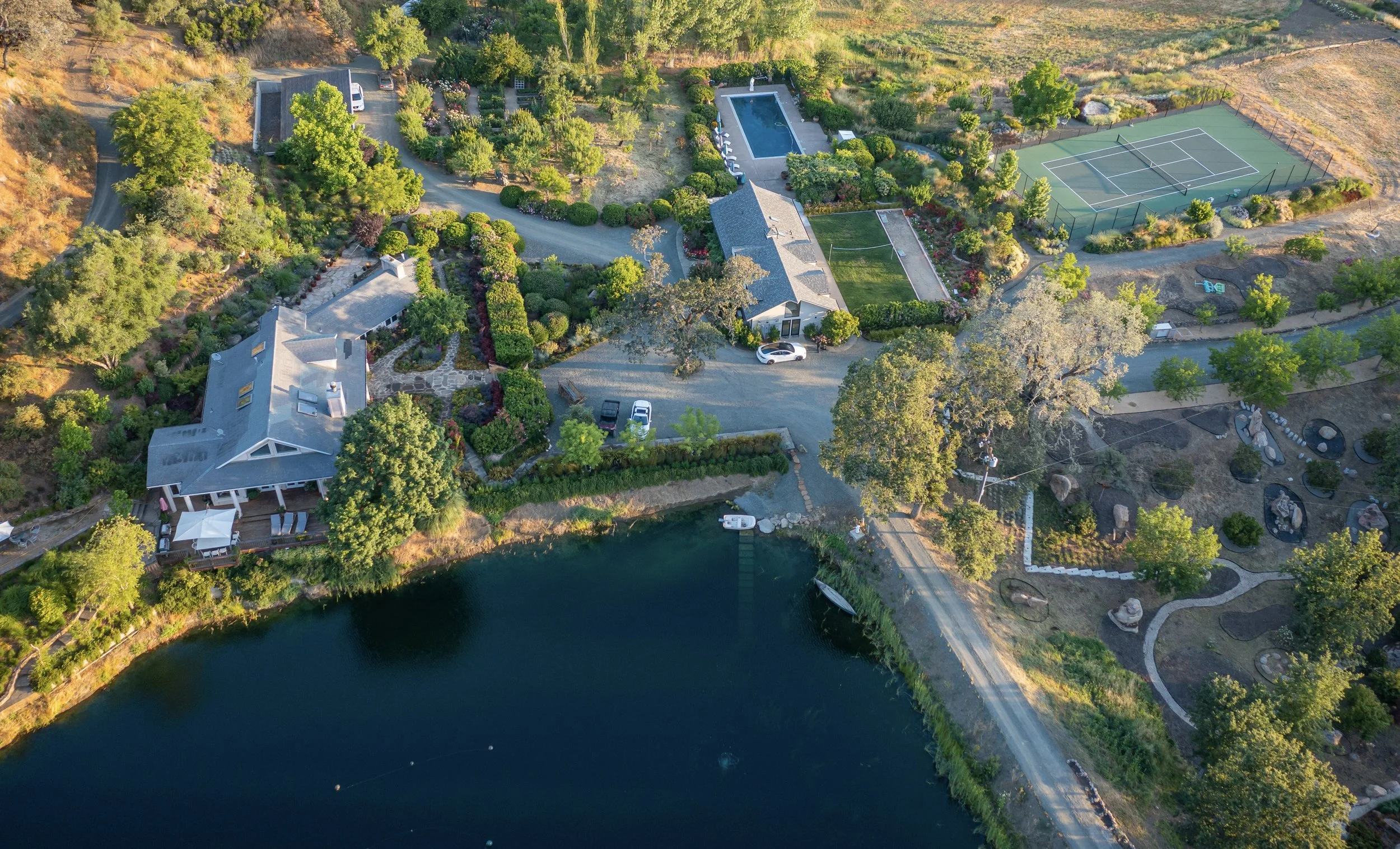 Aerial view of a spacious lakeside property with a house, swimming pool, tennis courts, lush gardens, and a parking area surrounded by trees.