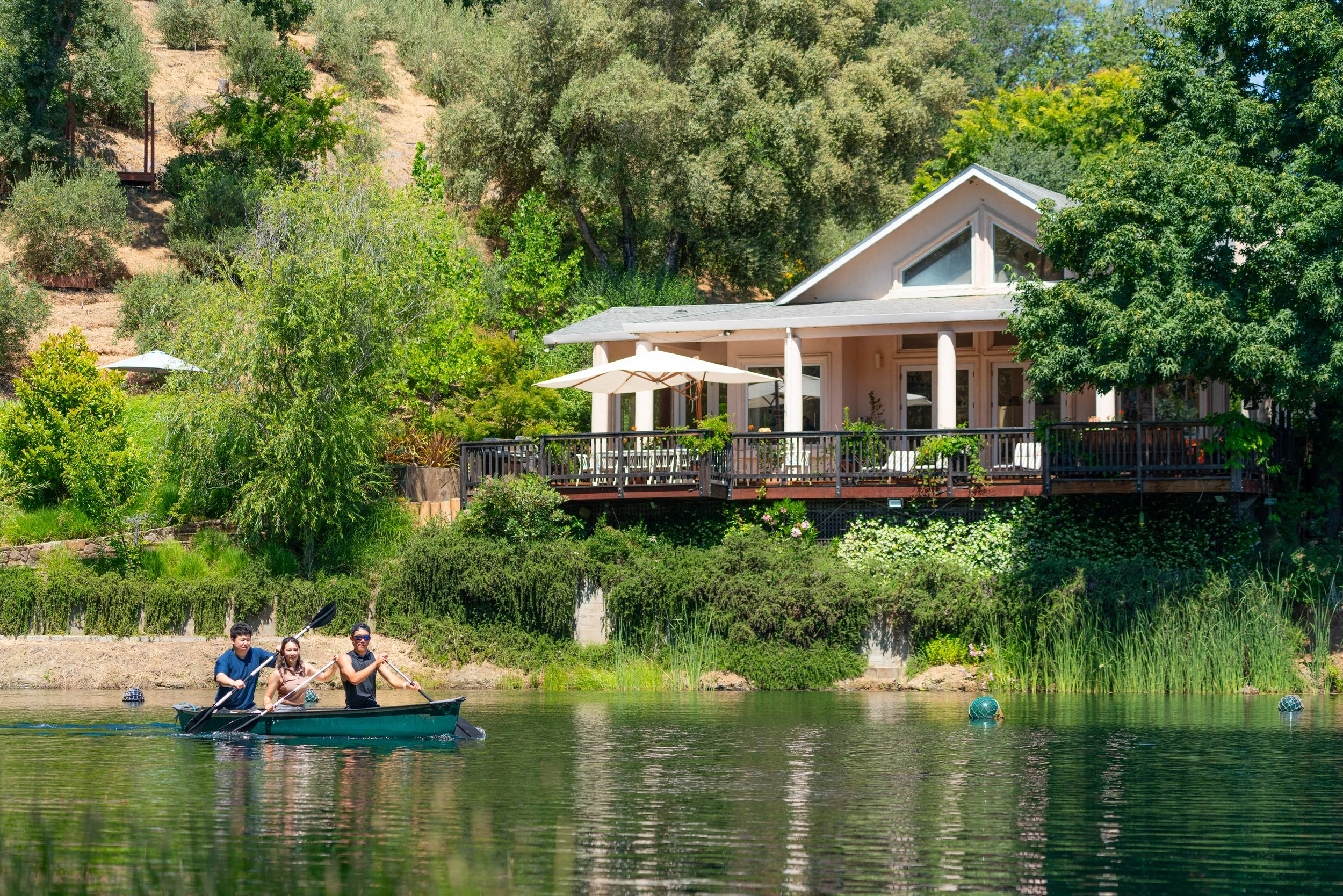 Three people are paddling a canoe on a lake in front of a house with a large deck, surrounded by lush green trees and plants.