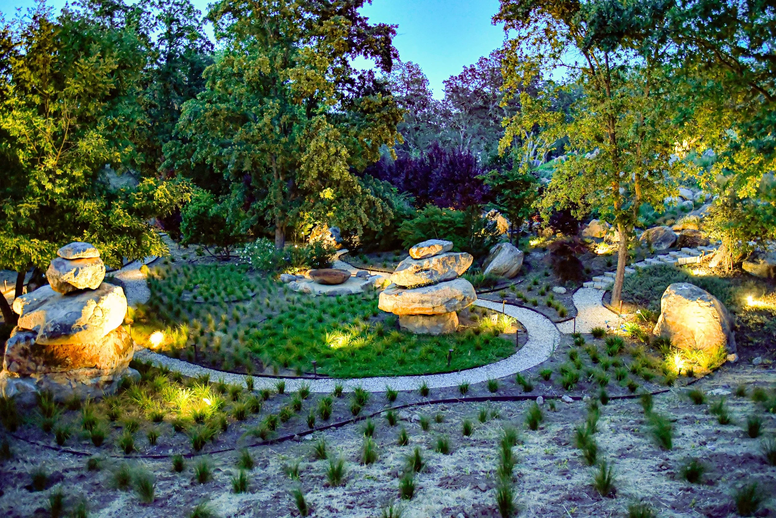 A curated garden at dusk with stone sculptures, a winding pebble pathway, various trees, and landscaped plants illuminated by outdoor lights.