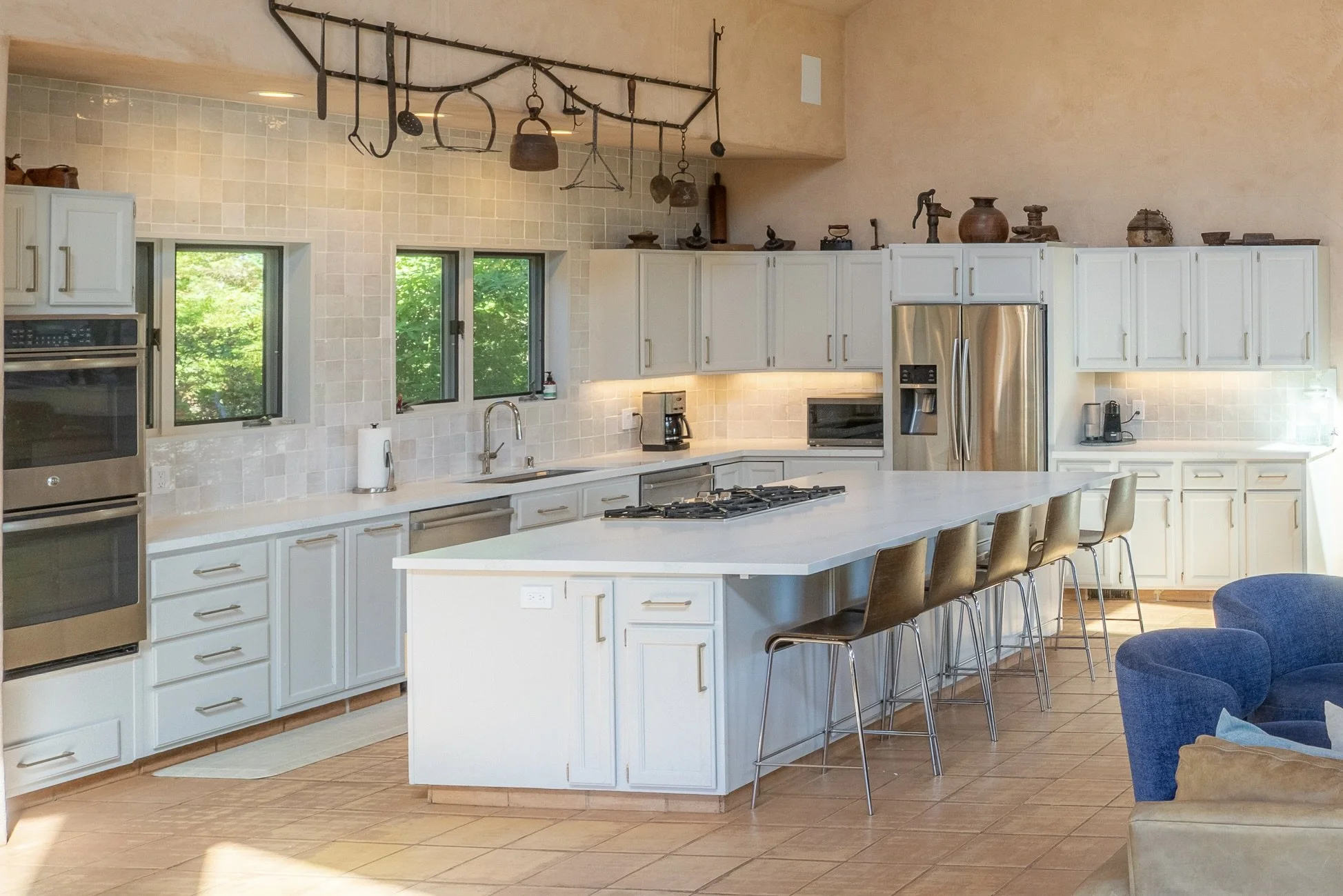 Modern kitchen with white cabinets, stainless steel appliances, a large island with seating, and a tiled backsplash, with windows overlooking green trees.