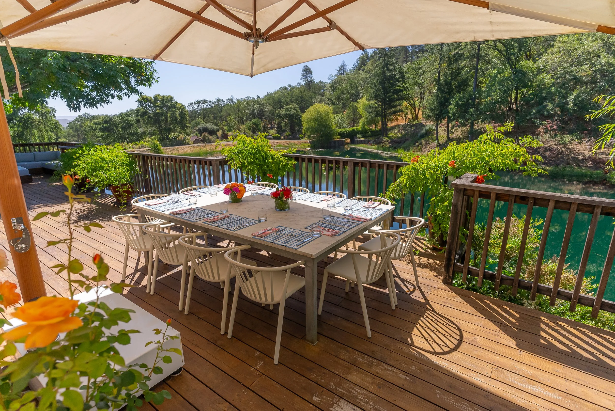 Outdoor dining table set with eight chairs, paper placemats, glasses, and floral centerpieces on a wooden deck overlooking a lake, shaded by a large umbrella with trees and hills in the background.