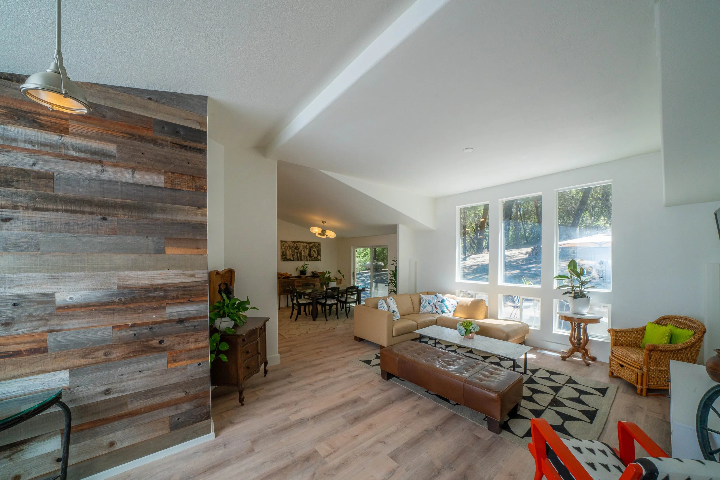 Bright living room with large windows, cream sectional sofa, patterned pillows, wooden side tables, indoor plant, and a brown leather ottoman on a geometric rug.