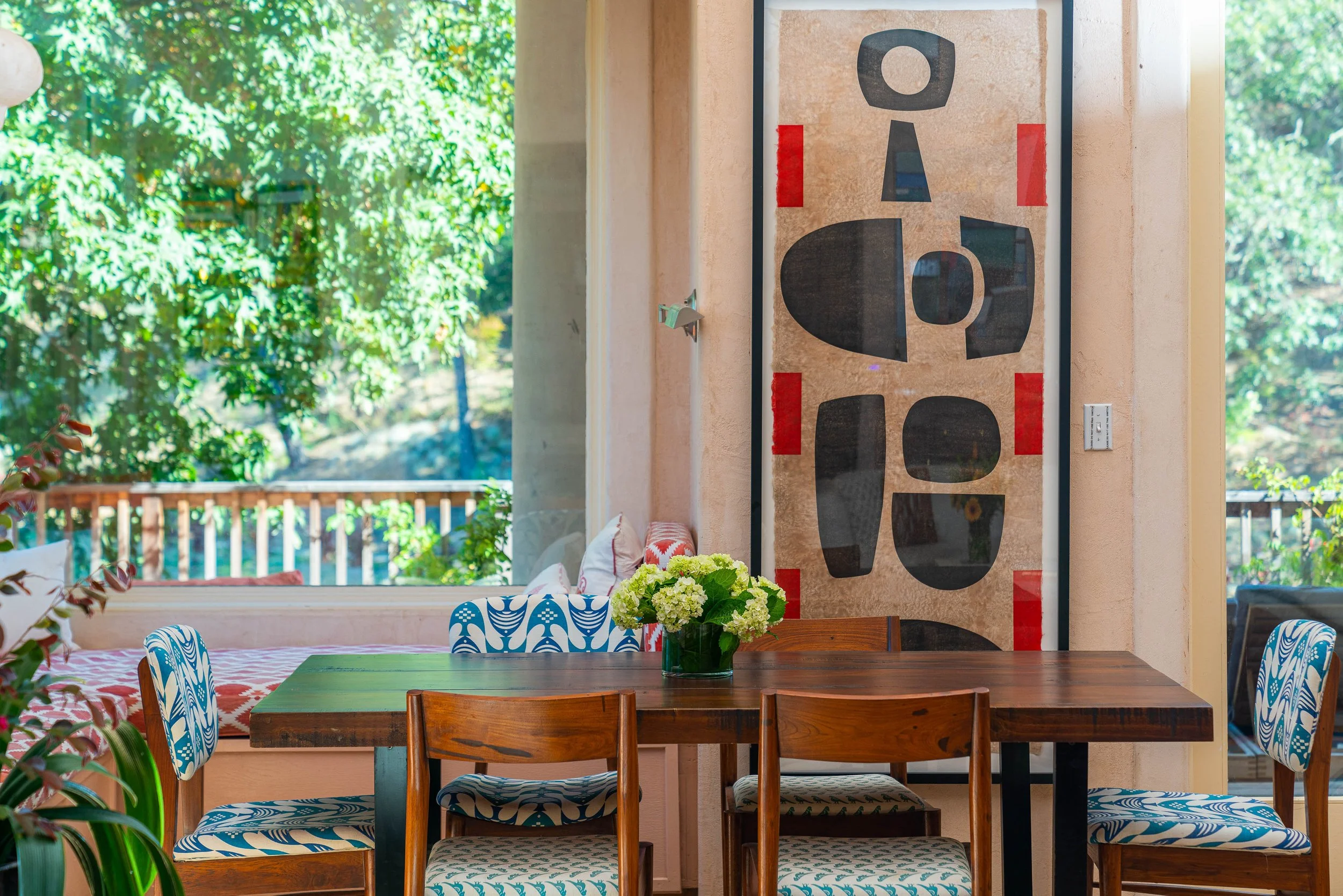 Interior dining area with a wooden table, blue and white patterned chairs, a vase of white flowers, and large window showing green trees outside.