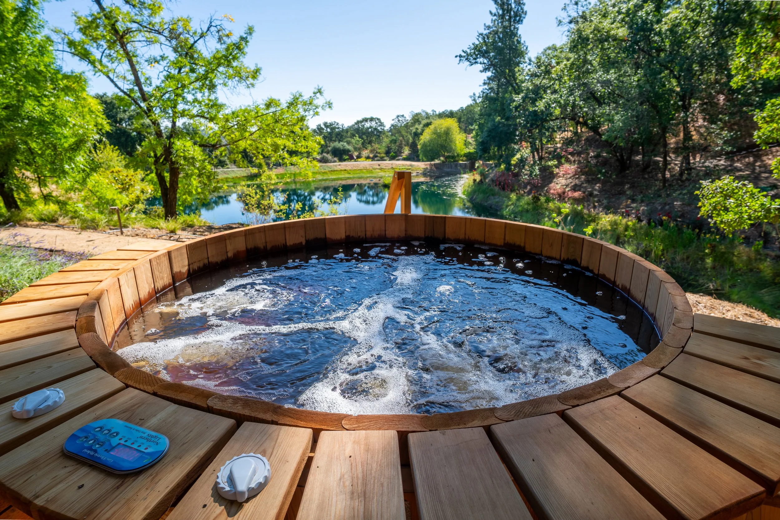 Hot tub outdoors overlooking a lake and lush green trees on a sunny day.
