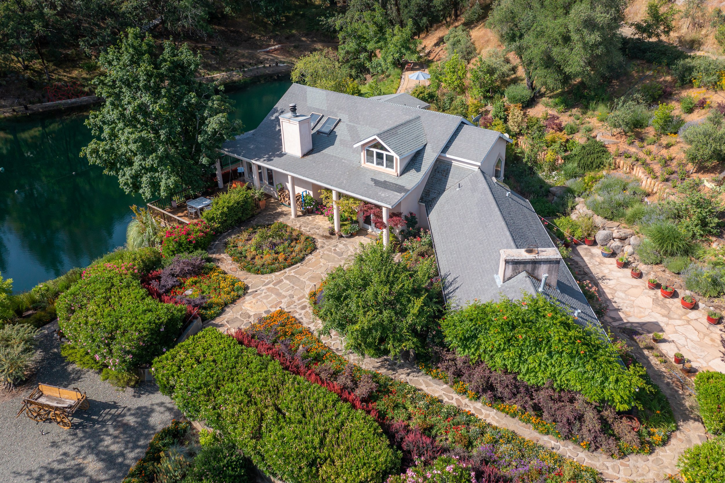 A large house surrounded by colorful garden beds and trees nestled next to a pond or lake, with a stone pathway leading to the front porch and a patio area on the side.