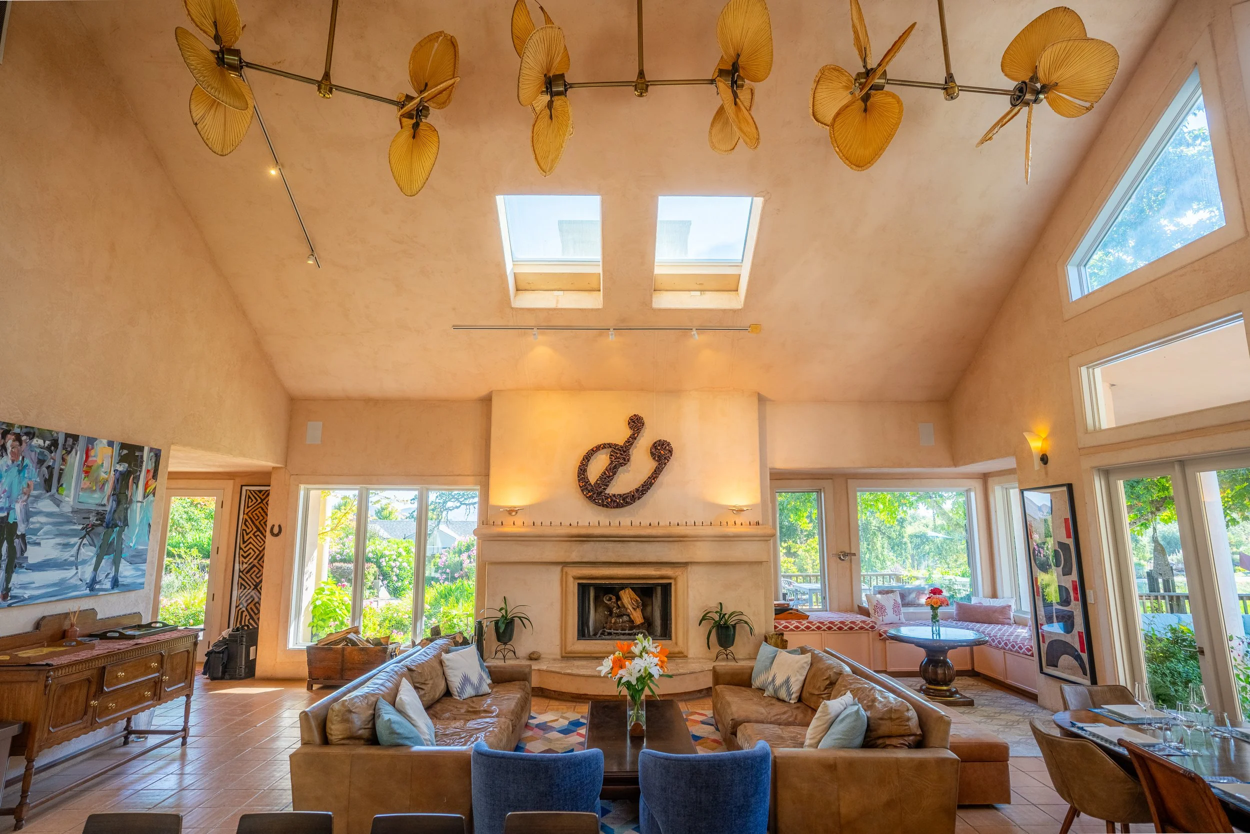 Living room with a central fireplace, two brown leather sofas with white pillows, a wooden coffee table with a flower arrangement, artworks, windows showing green outdoor scenery, and a unique floral ceiling light fixture.