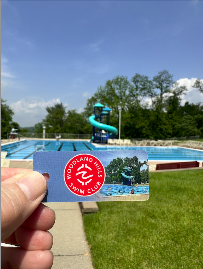 A person holding a Woodlands Hills Swim Club membership card in front of a swimming pool with a water slide in the background during daytime.