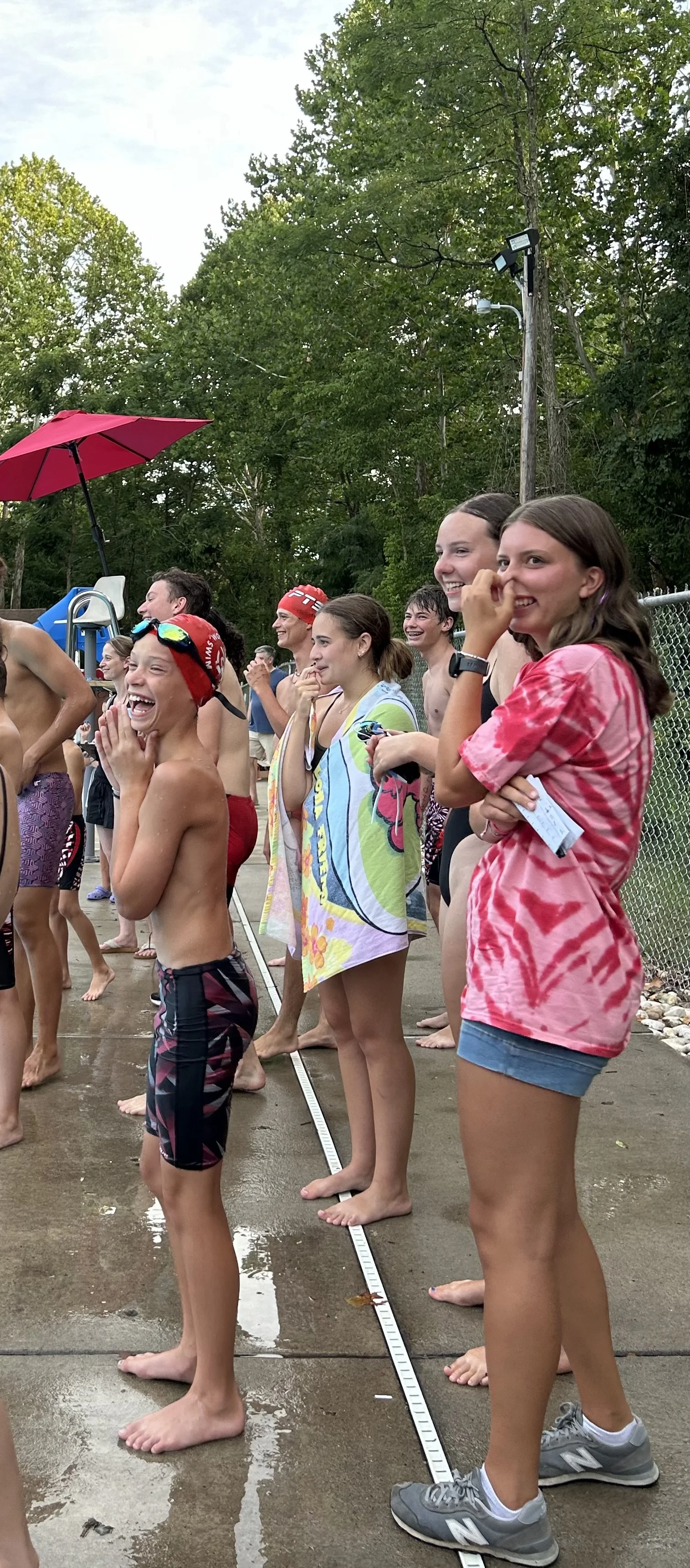 Group of young people at a water park, some in swimsuits and others in casual clothes, smiling and laughing. A girl in the foreground is wearing a pink tie-dye T-shirt and shorts, standing on wet pavement near a pool area, with greenery and a fence in the background.