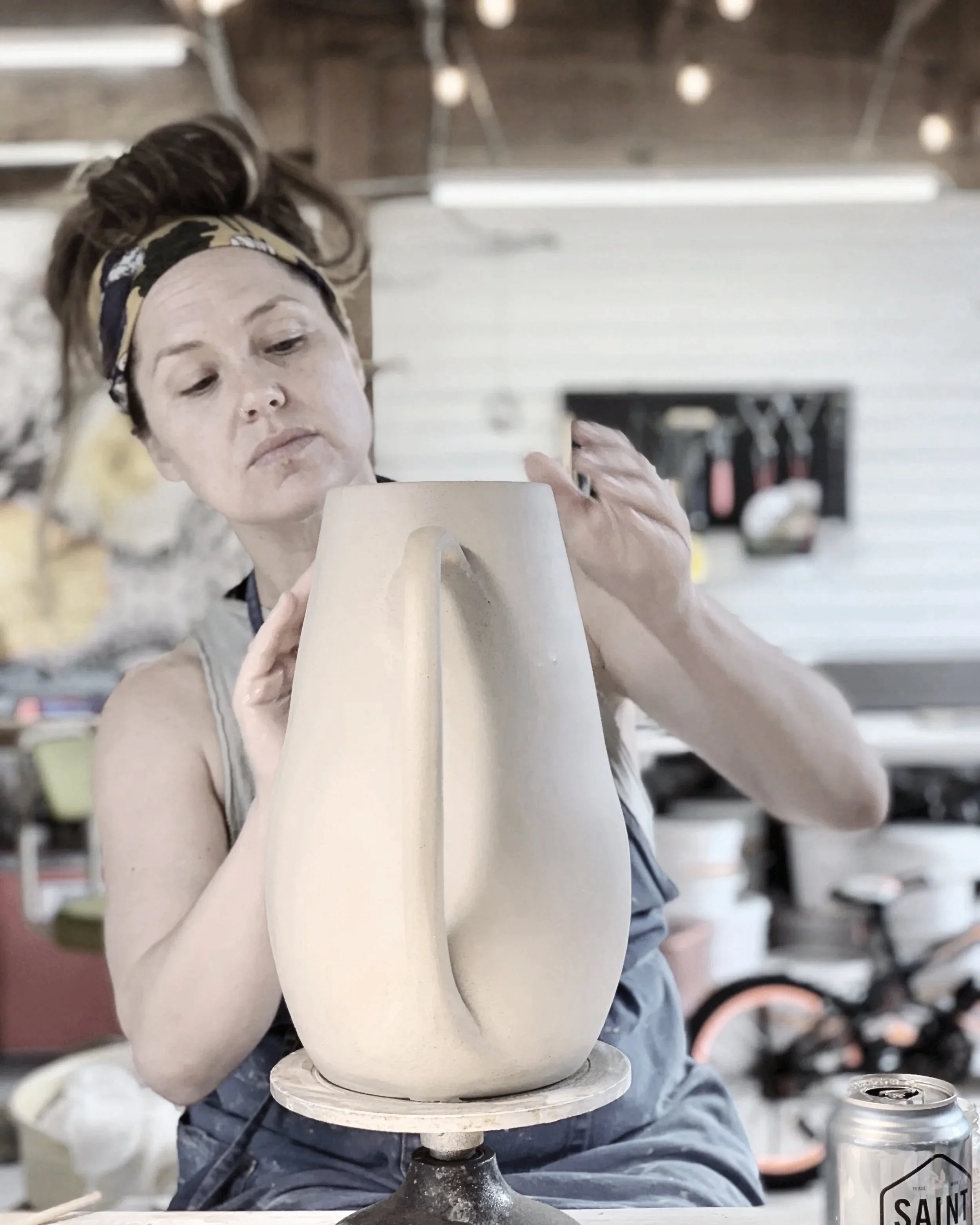 A woman in a workshop wearing a headband, shaping a piece of clay on a spinning pottery wheel.