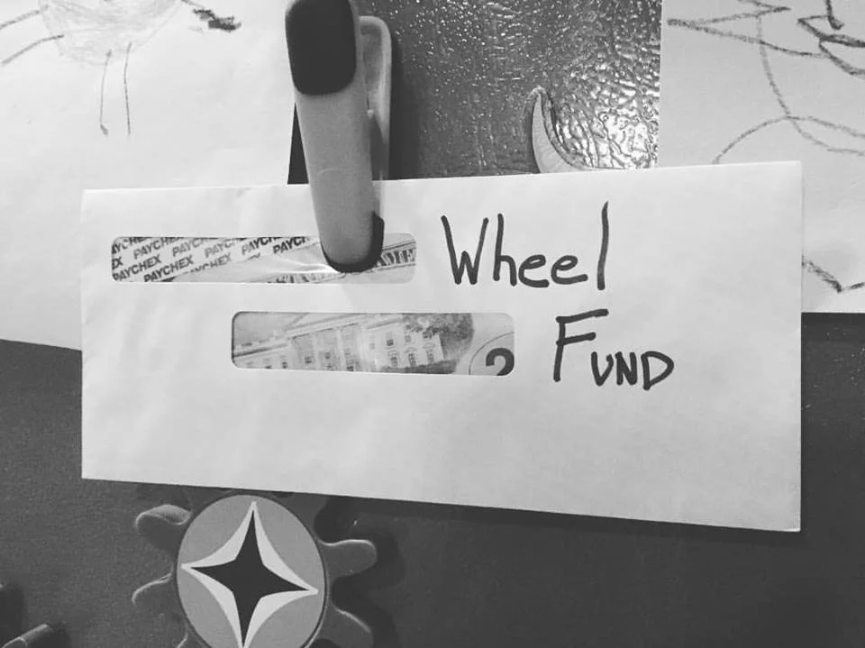 A paper labeled 'Wheel Fund' held with a clip over a desk. A Pittsburgh Steelers logo pen is attached at the bottom of the paper, and a piece of tape with the word 'Paychex' written repeatedly is at the top.