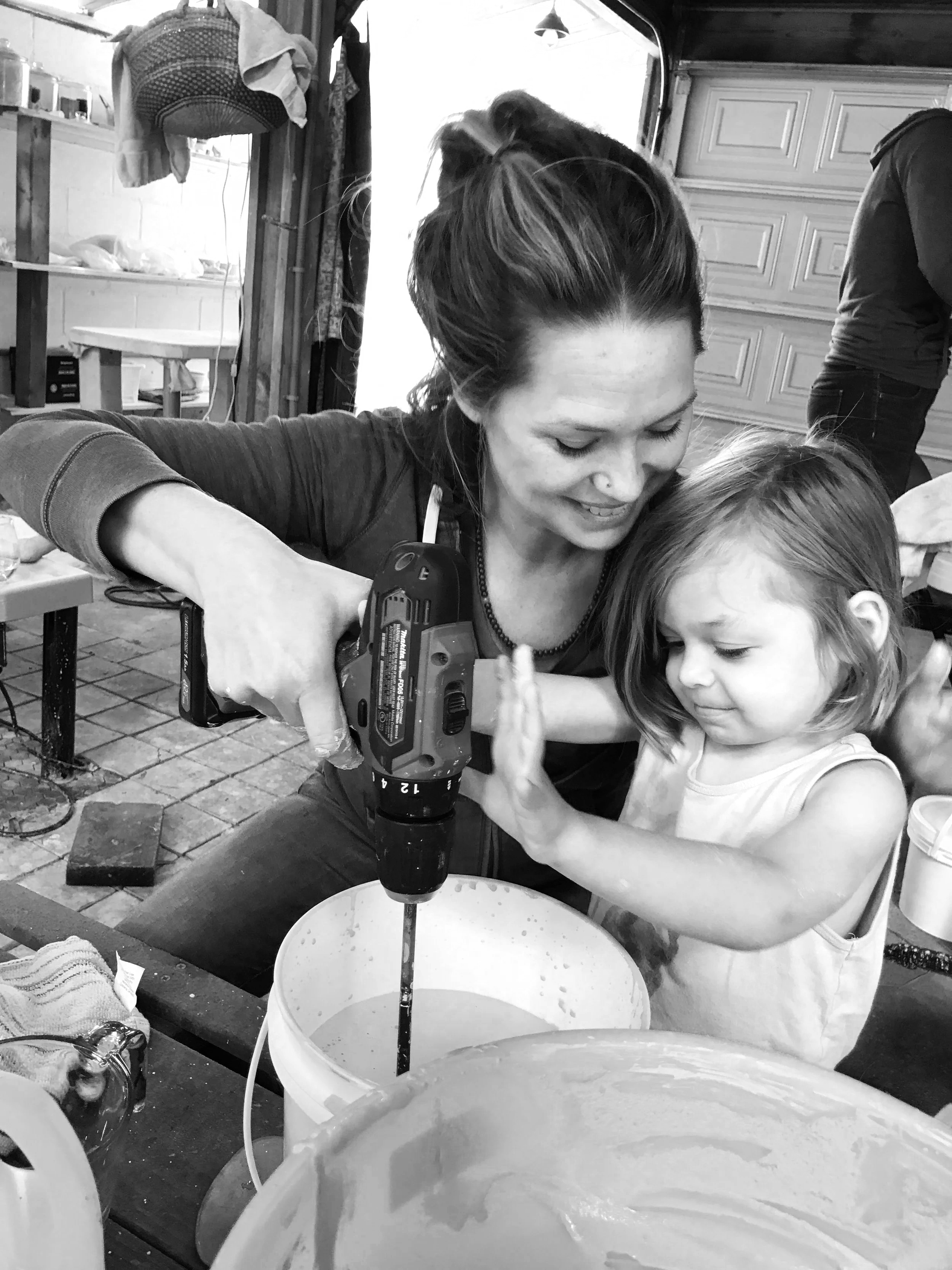 A woman and a young girl are working together to stir a large bowl of liquid using a power drill. They are in a workshop or kitchen area with various tools and supplies visible in the background. Both appear focused on their task, sharing a moment of teamwork and concentration.