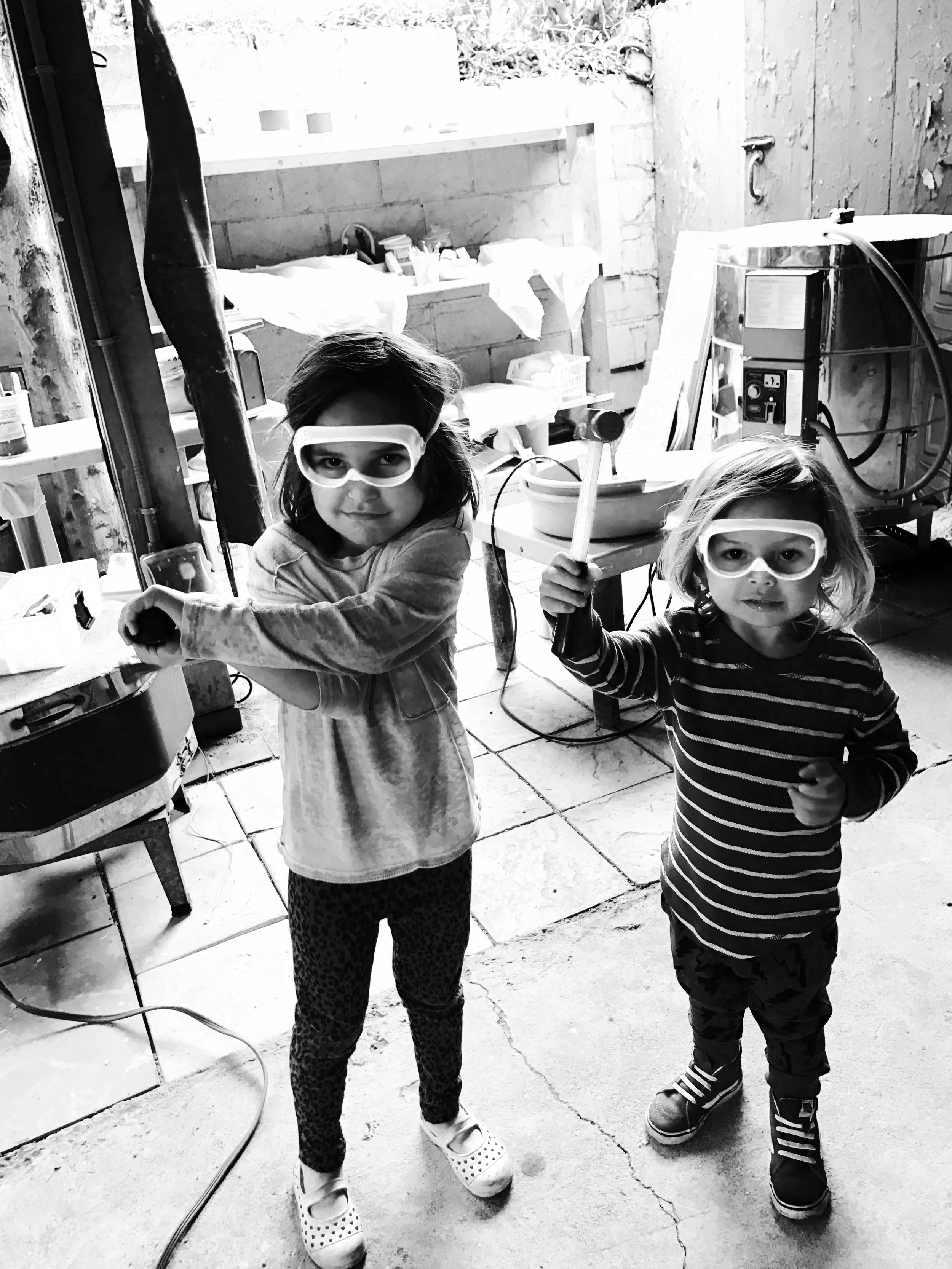 Two young children, one girl and one boy, wearing safety goggles, in a workshop or garage, holding tools, with workshop equipment and supplies in the background.