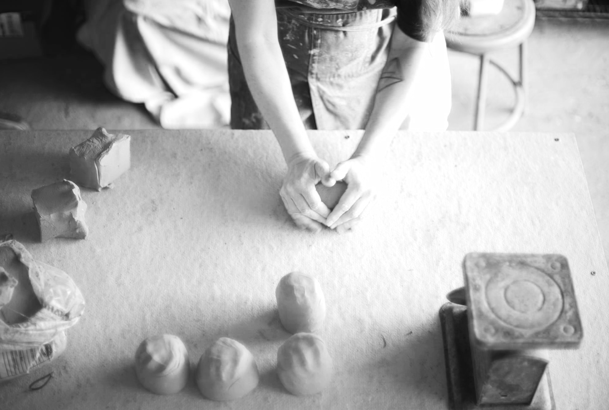 Black and white photo of a person making a heart shape with their hands over a table with clay objects.