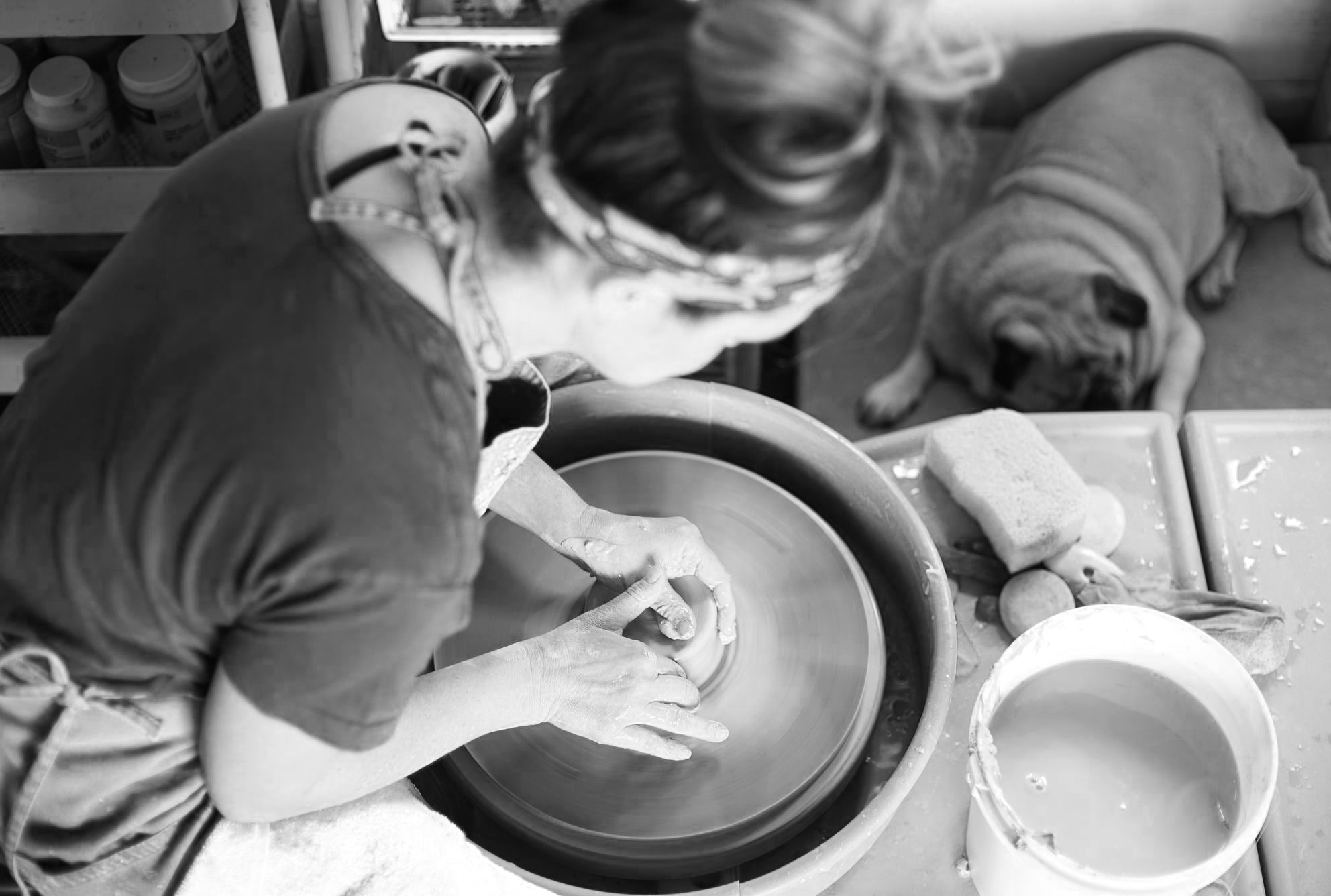 A person shaping clay on a pottery wheel with a dog lying nearby, surrounded by pottery tools and supplies.
