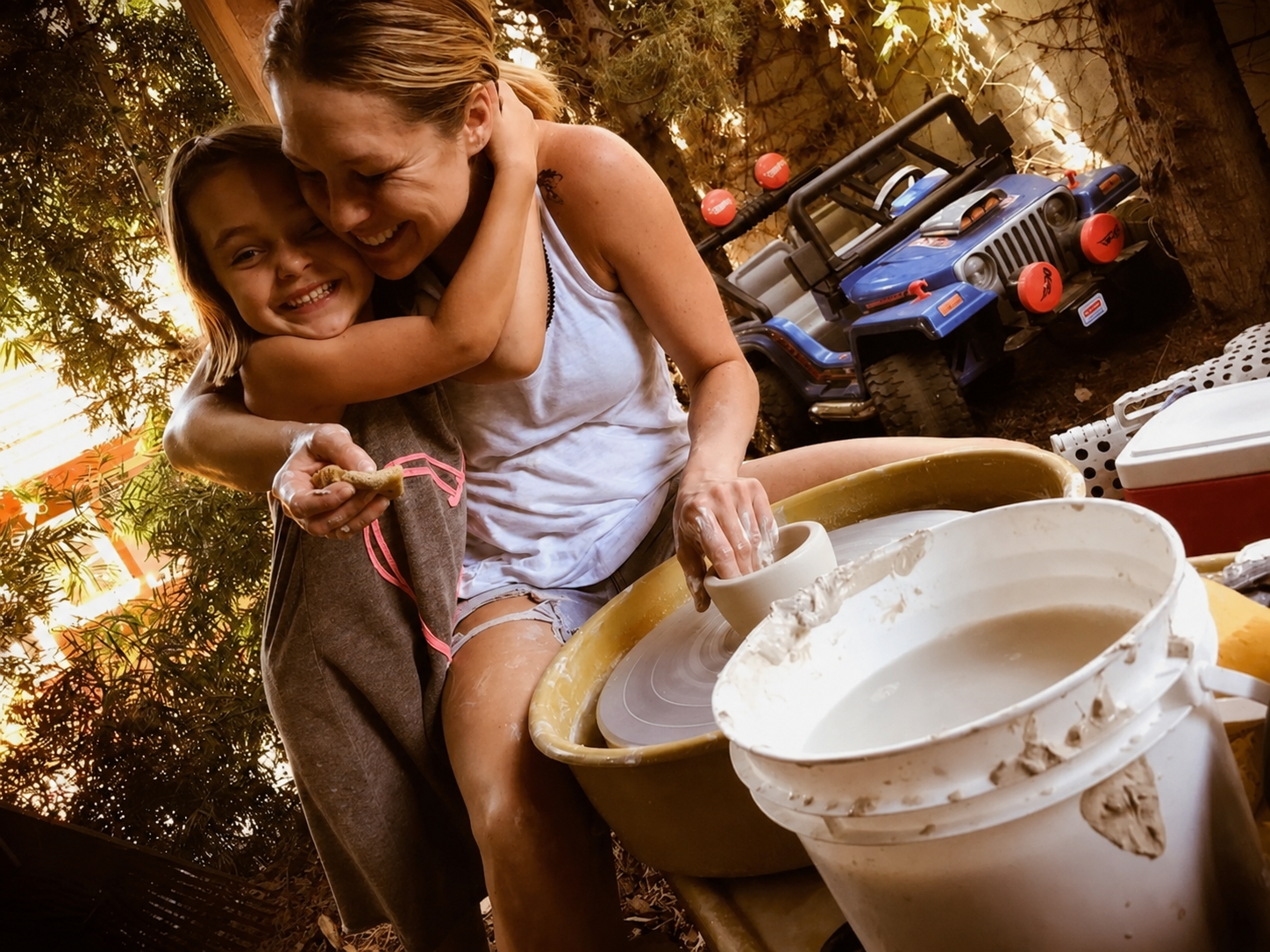 A woman and a young girl are laughing and hugging as the woman molds clay on a pottery wheel outdoors. There are buckets of water, a red and white cooler, and toy vehicles in the background amidst trees and warm lighting.